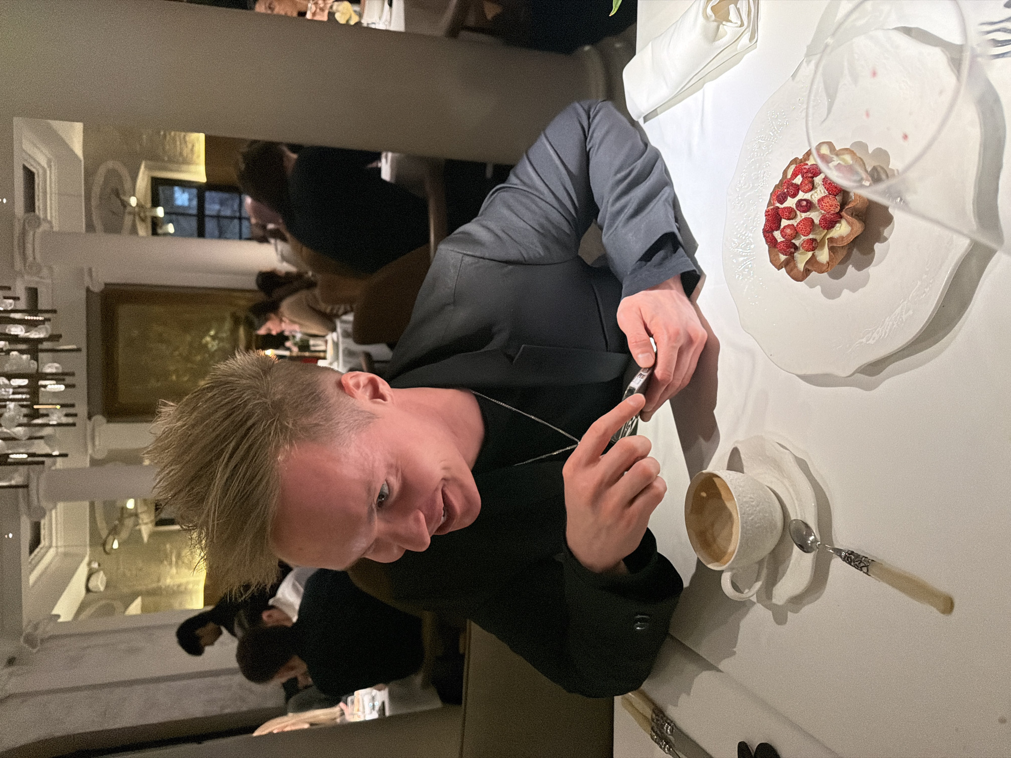 A fair-skinned man with blonde hair is taking a photo of his food at a restaurant. He is wearing a black blazer and a silver necklace. A plate with a strawberry tart and a coffee cup with a spoon is on the table in front of him. Other people are seated at tables nearby, and there is a decorative chandelier in the background. Based on the ornate interior, this scene could be taking place in a high-end restaurant, possibly in Moscow, Russia.