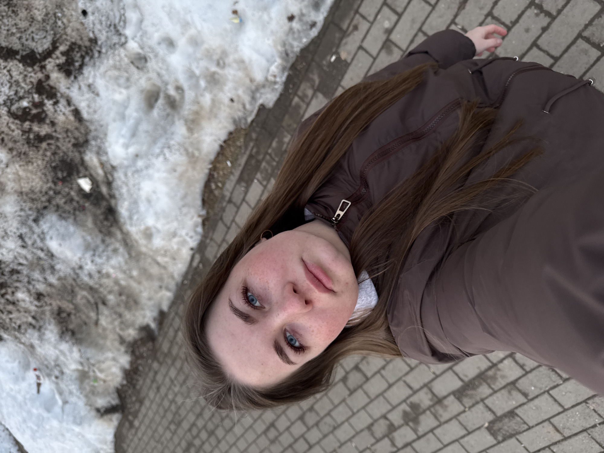 A young woman with light brown hair and blue eyes is seen in a tilted selfie. She is wearing a brown puffy jacket. The background includes melting snow on one side and a brick sidewalk on the other. The location is identified as the city of Chaikovsky, Russia.