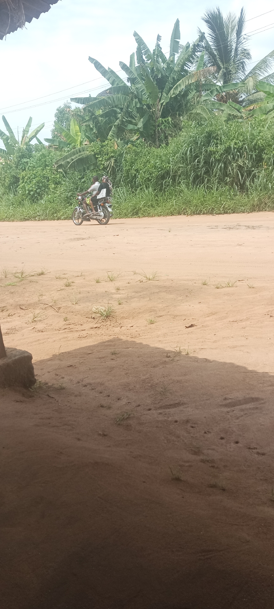 The image shows a dirt road in Amauzu, Nigeria. A motorcycle with two people is traveling on the road. The environment is tropical, with banana trees and other lush vegetation. The foreground is shaded, suggesting the photo was taken from a building or structure.