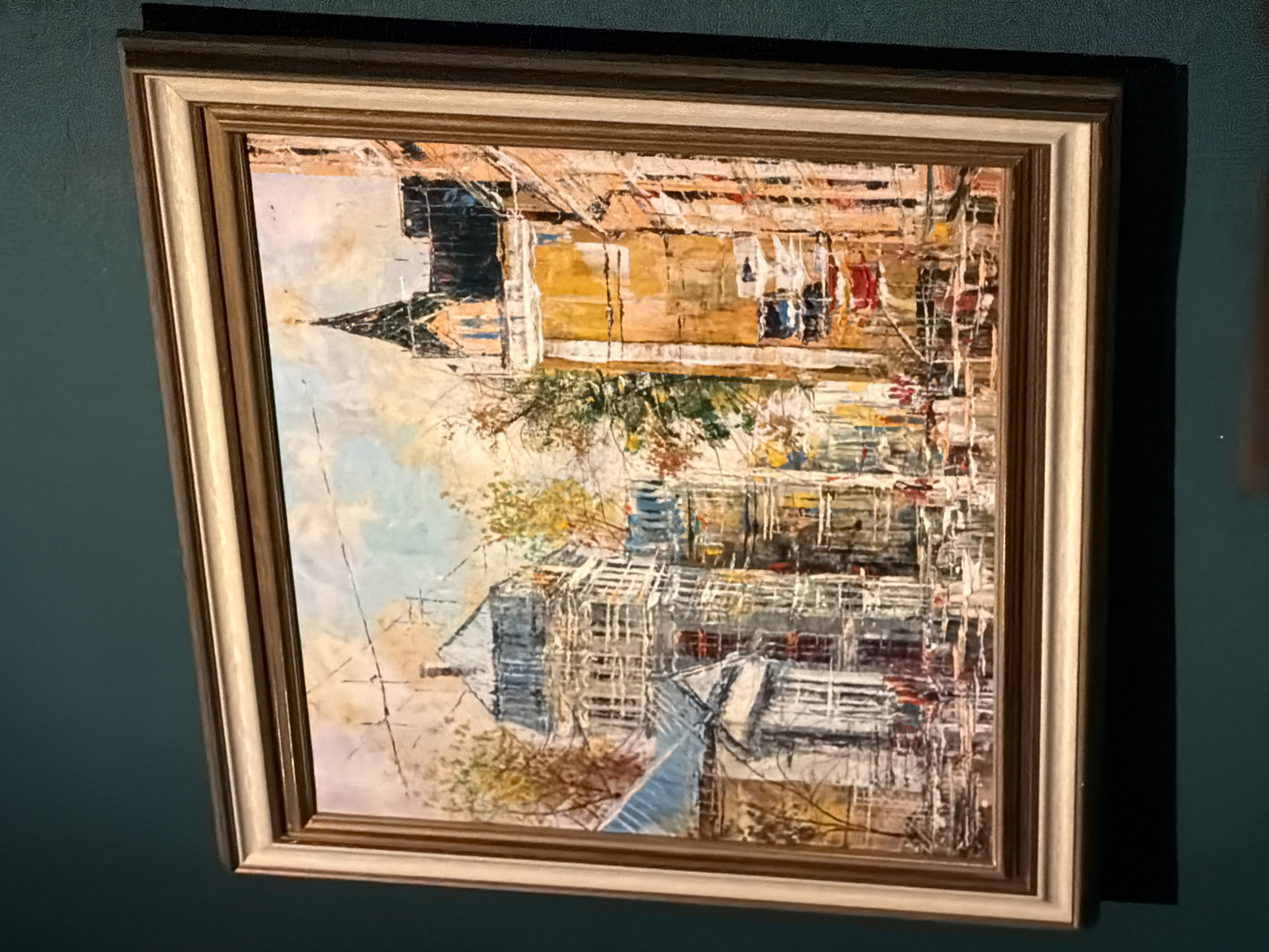 A framed painting hangs on a textured, dark teal wall. The artwork itself is an impressionistic or abstract cityscape, dominated by visible, textured brushstrokes. The central subject features buildings rendered in shades of yellow, brown, white, and blue, with accents of red and hints of greenery, possibly depicting trees with autumn foliage. Some sections of the structures have a layered or scaffold-like appearance. The painting is oriented vertically within its frame, portraying the buildings upright.
The frame surrounding the painting is made of light-colored wood, possibly gilded or a natural wood tone, and features a multi-tiered profile. A distinct shadow is cast on the wall to the right of the painting, indicating the direction of the light source. The wall surface itself has a visible, subtle texture. There is no discernible text present on the painting, frame, or wall.