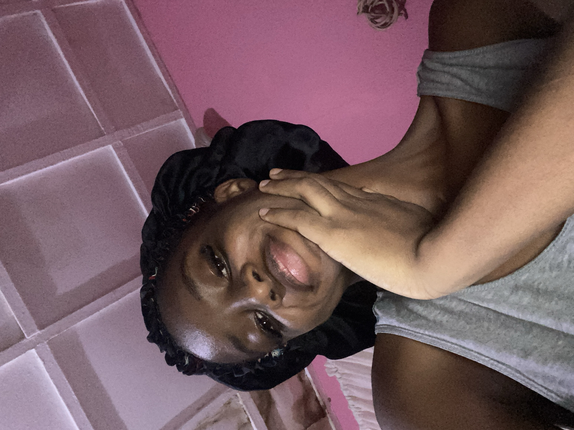 A young woman wearing a gray tank top and a black satin bonnet is looking at the camera with her hand on her chin. Her eyes are wide and she has a slight smile. The background is a pink wall and a white ceiling with visible light fixtures. The scene appears to be indoors, likely in a bedroom, and the lighting suggests it might be nighttime or a dimly lit room. The image quality is adequate, with a slight graininess. There is no indication of the location being in Jos, Nigeria.
