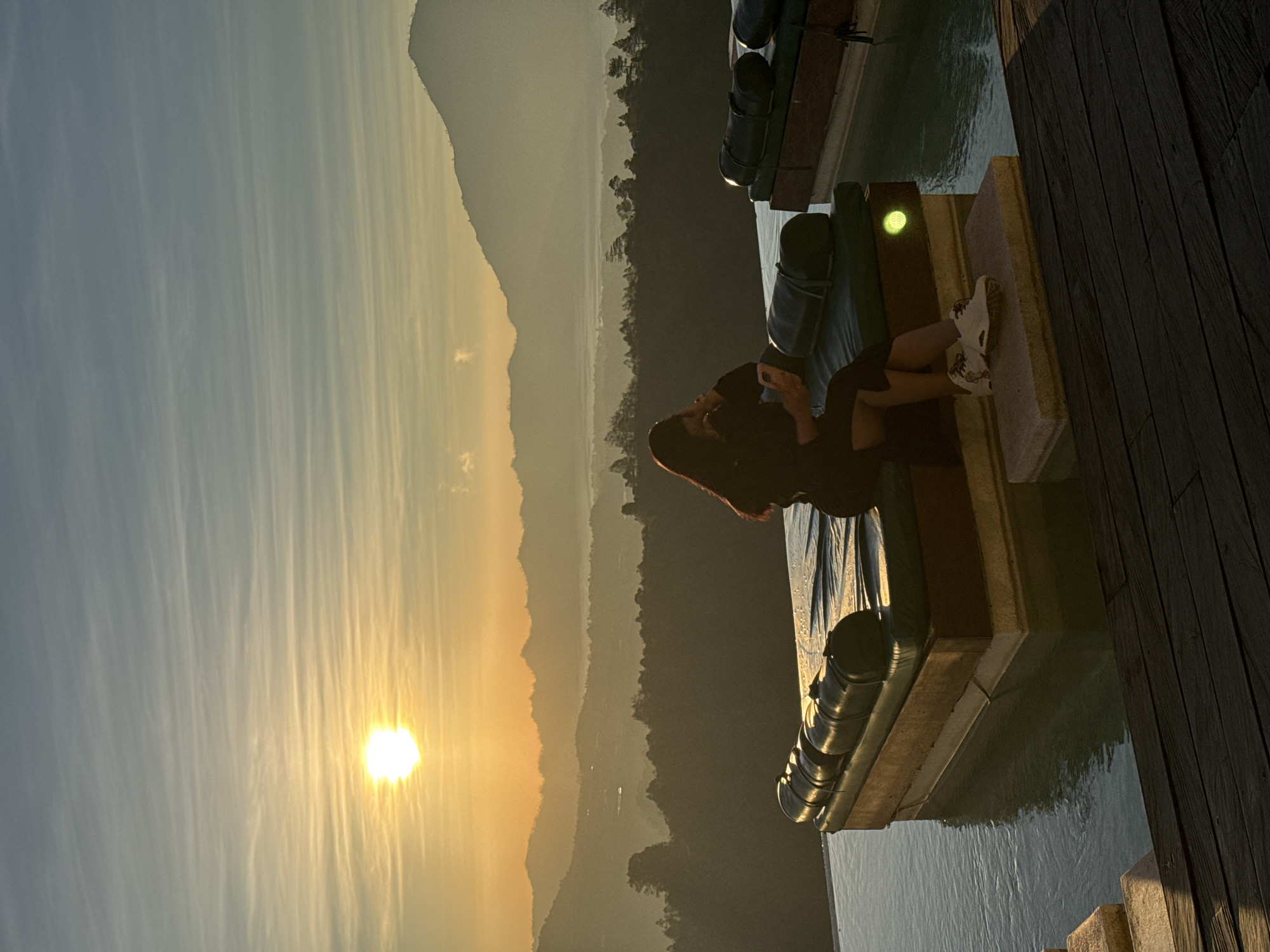A woman is depicted sitting on a submerged lounge chair in an infinity pool, observing a sunrise or sunset over the mountainous landscape of Kintamani, Indonesia. The scene is illuminated by the low-lying sun, positioned in the upper left quadrant, casting a golden hue across the sky and distant terrain.
In the foreground, a dark wooden deck occupies the bottom right and lower edge of the frame. Two light-colored stone steps or platforms extend from the deck into the infinity pool. The pool water, a clear blue-green, reflects the ambient light. Two dark green lounge chairs, each with a rounded bolster cushion, are situated on stone bases partially submerged in the pool. A small, circular, yellow-green light fixture is visible on the side of the lounge chair base closer to the viewer.
The sole person, a woman, is seated on the left lounge chair. Her back and right side are visible as she faces away from the viewer, looking towards the horizon. She has long, dark hair and is wearing a dark-colored dress or skirt with a slit on the left side, revealing her leg. She wears white athletic shoes with dark intricate patterns. Her left hand rests on her lap, holding a light-colored object, likely a smartphone. Her right hand is raised near her head, suggesting a pensive pose or an adjustment of her hair.
The midground reveals multiple layers of forested hills and mountains, receding into the distance. A distinct layer of atmospheric haze or mist lies in the valleys and between the mountain ranges, particularly noticeable in the center and right midground.
The background features additional mountain peaks, including a prominent, larger mountain silhouetted on the far right. The sky above is gradient, transitioning from a bright golden at the horizon to a lighter blue-grey overhead. Wispy, horizontally oriented clouds are scattered across the upper sky, catching the warm light from the sun.