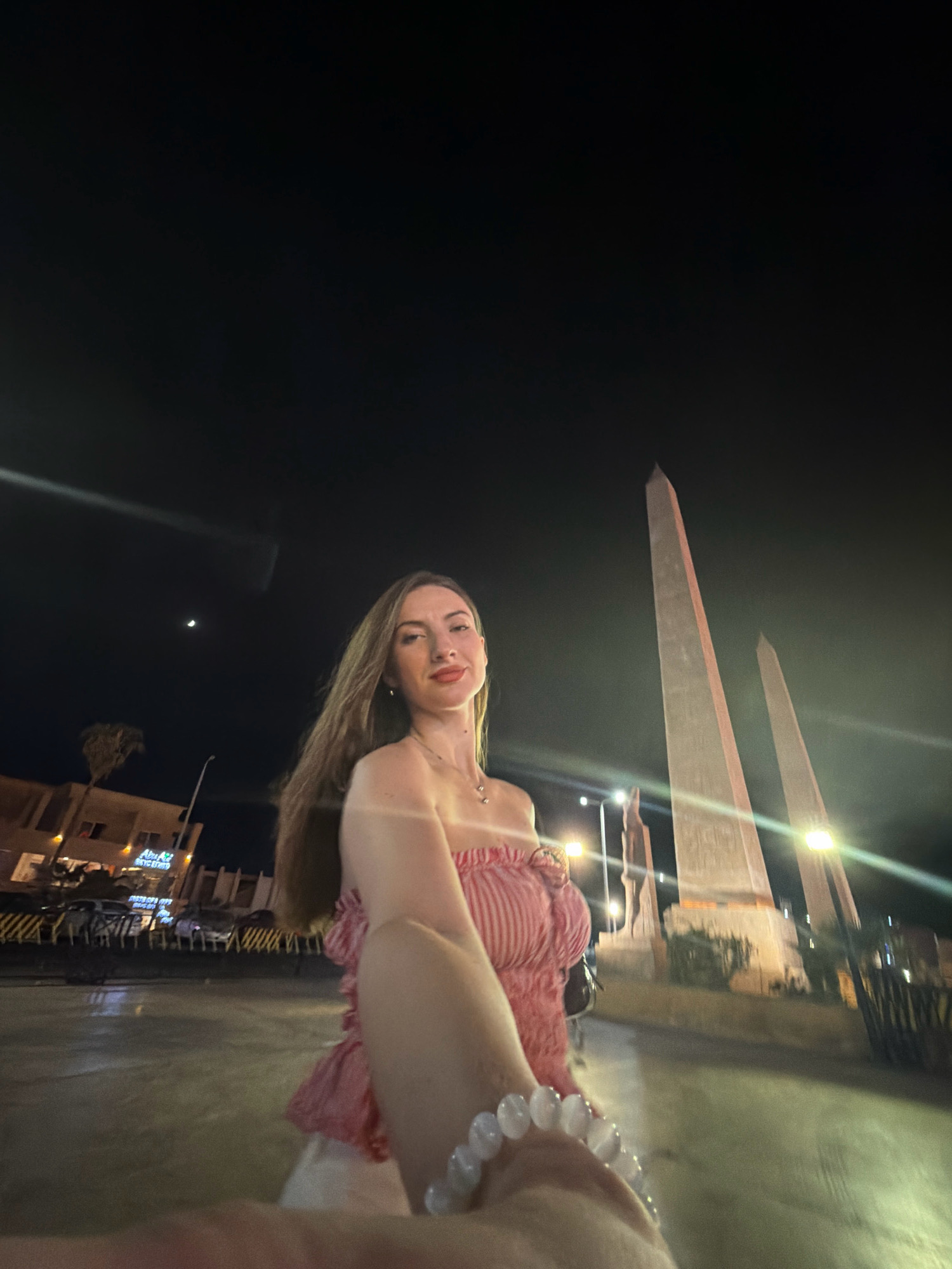 A woman with long brown hair and red lipstick is taking a selfie at night in Sharm El Sheikh, Egypt. She is wearing a strapless red and white striped top and white pants, and a beaded bracelet on her wrist. In the background, two tall obelisks are illuminated, and there are streetlights and buildings. A crescent moon is visible in the dark sky. The ground appears wet, possibly from rain or a sprinkler system.