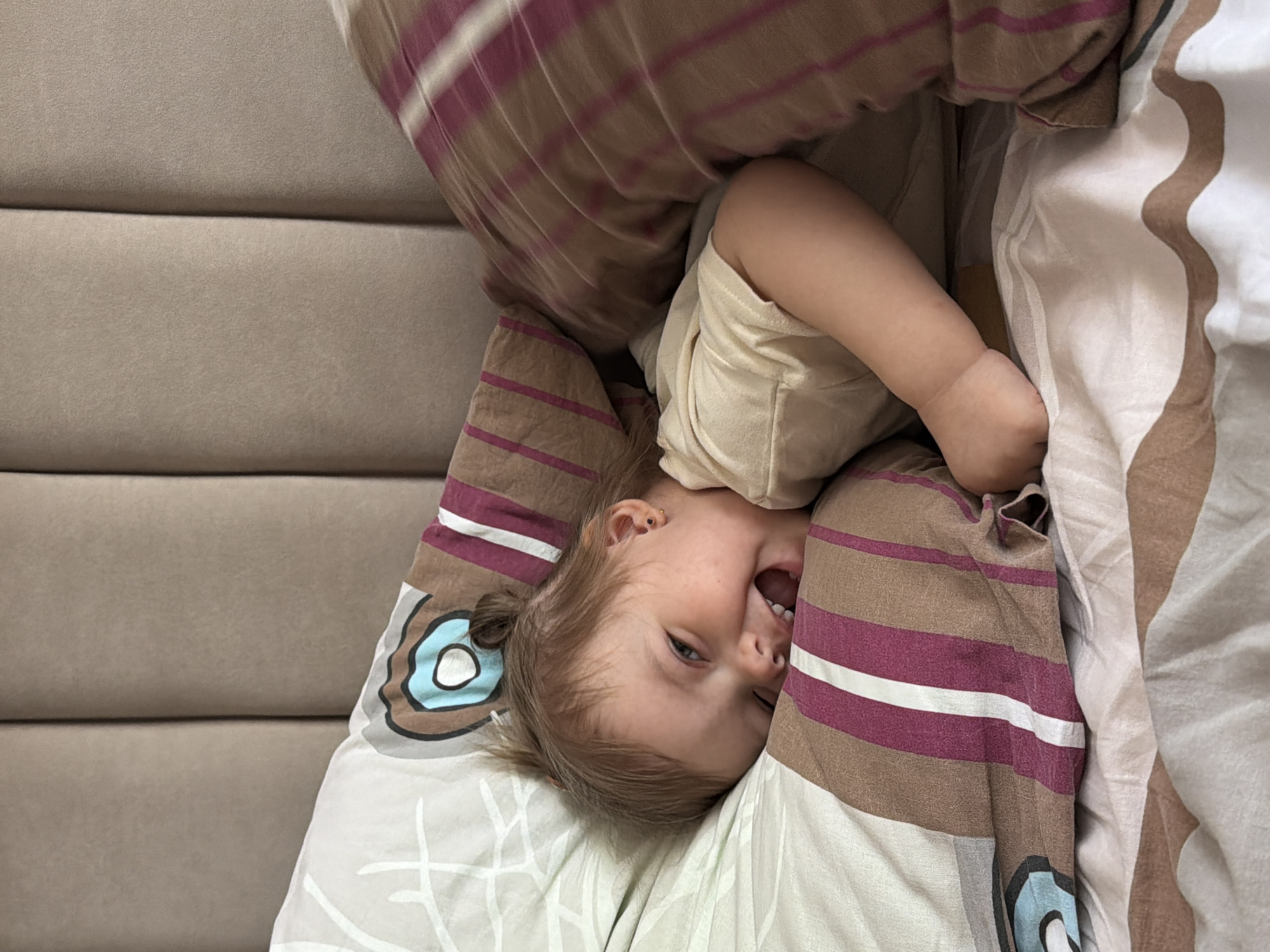 A child is lying on a bed with a pillow and duvet. The child is positioned upside down with their head near the bottom of the frame. The bedding has a striped pattern with brown, white, and pink lines. The pillow has a blue donut-shaped design. The bed appears to be located indoors. There is no information to confirm the location is in the city of Reshetnikovo, Russia.