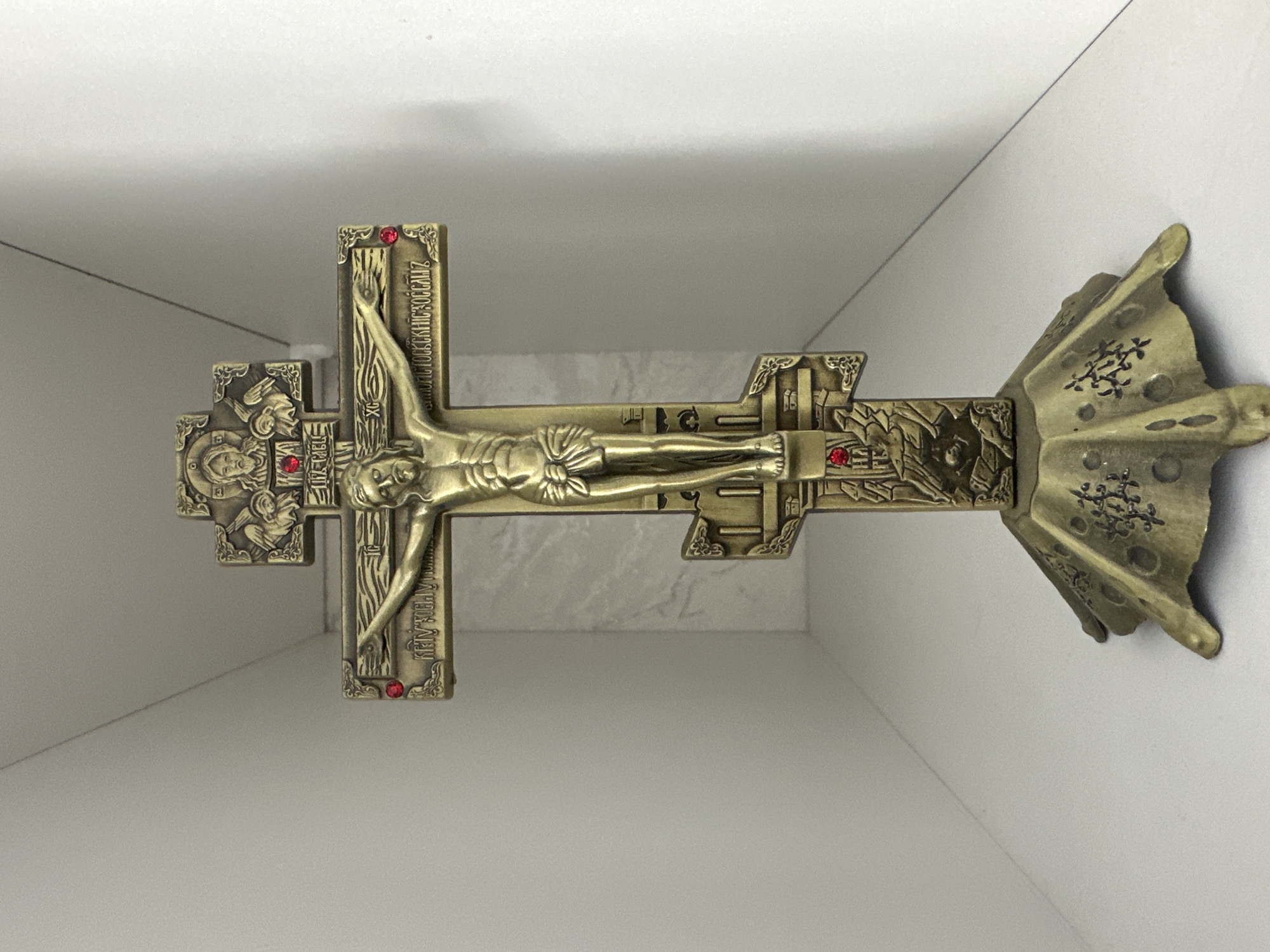 This is a media file depicting a bronze-colored crucifix with intricate details. The crucifix features a representation of Jesus Christ, with his arms outstretched on the cross. Inscriptions in Cyrillic script are visible on the crossbars, though they are difficult to fully decipher. Small red stones are embedded at various points on the crucifix, adding decorative accents. At the base of the crucifix, a stylized, fan-like element is present, also adorned with decorative carvings. The crucifix is presented against a plain, light-colored background, suggesting it is either displayed or stored in a simple setting. The lighting is even and appears to be artificial, indicating an indoor environment. There are no overt indications of a specific time of day, weather conditions, or emotional cues beyond the solemnity inherent in the religious imagery. The setting offers no distinct geographical clues beyond the provided location context of Zarinsk, Russia.