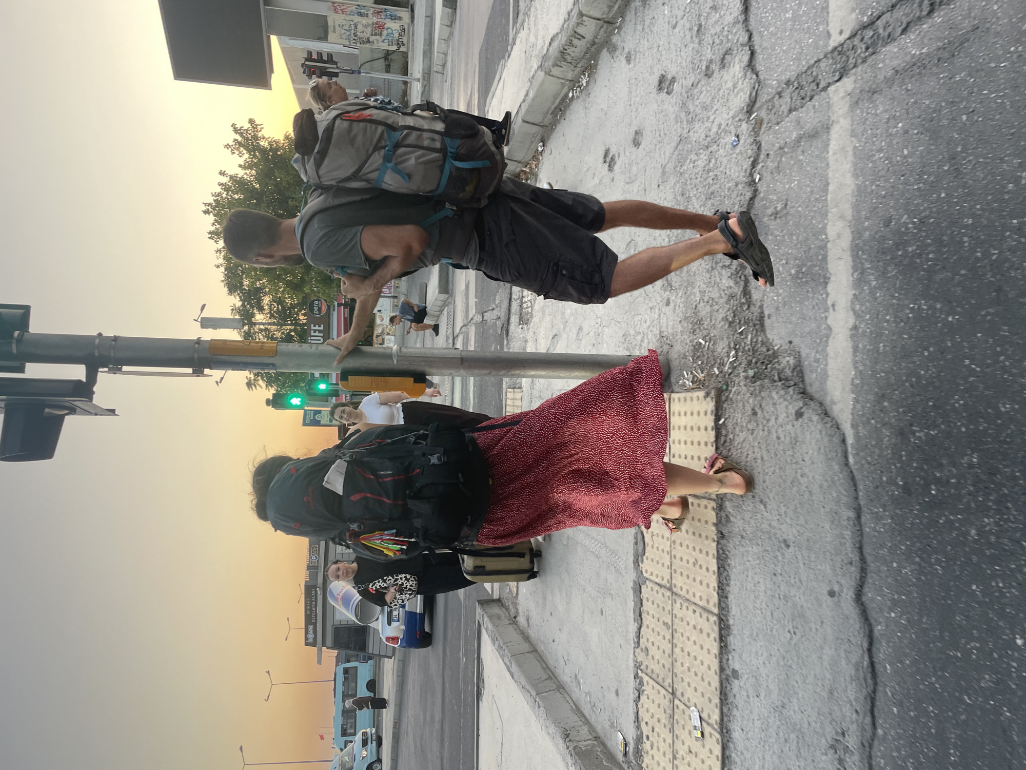 In Kadıköy, Turkey, at a pedestrian crossing, two individuals, a male and a female, stand near a traffic light pole. The male, wearing a grey t-shirt and cargo shorts, has a large grey backpack on his back and is leaning against the pole with his left hand. The female, with her back to the camera, wears a red polka-dot dress and has a large black backpack. A suitcase rests on the ground next to her. In the background, other people are visible, along with vehicles including a blue bus and a white car. The scene is illuminated by the warm glow of the setting sun.