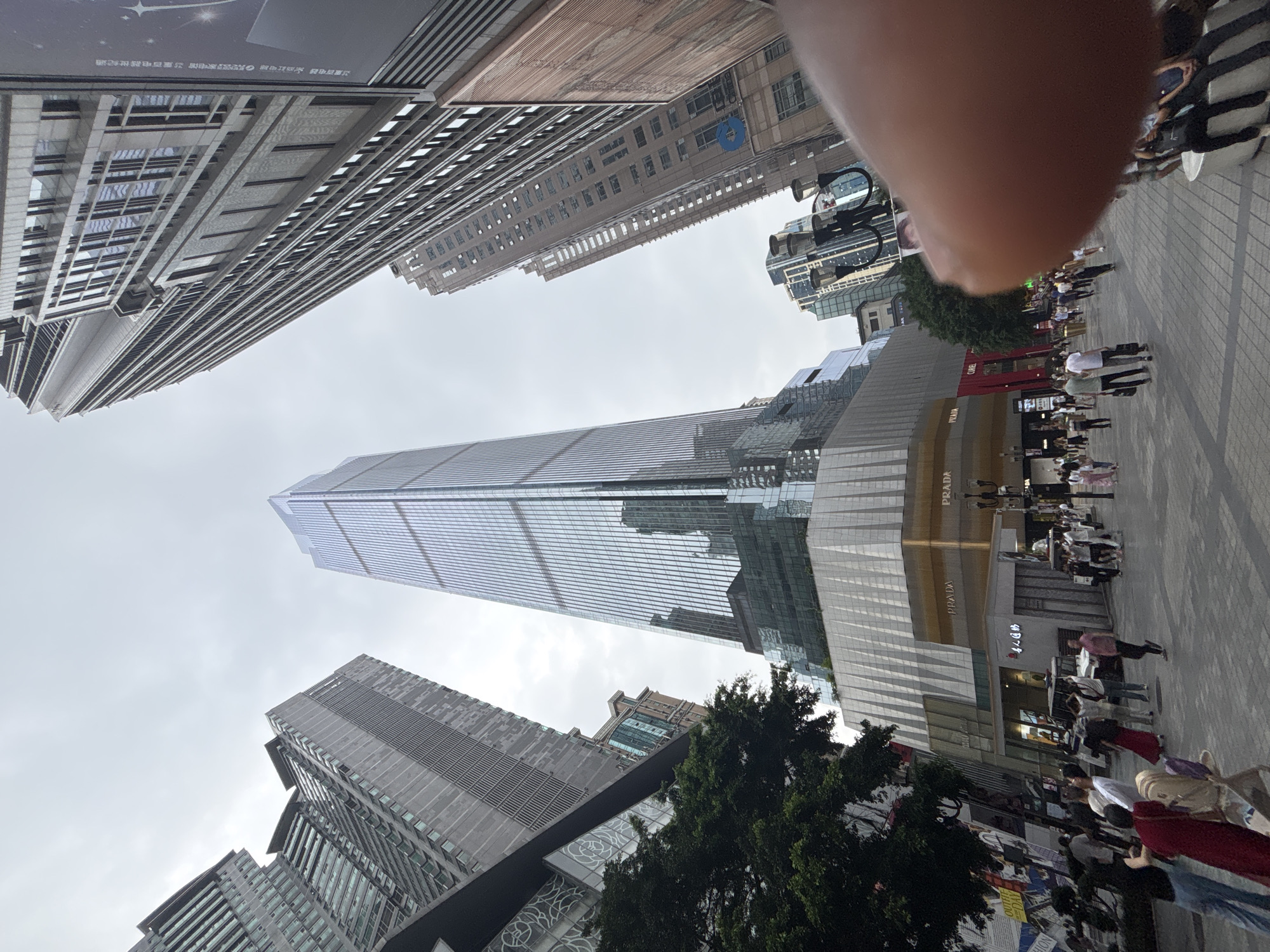 This image shows a street scene in Yuzhong District, Chongqing, China. The camera points upward, capturing the towering skyscrapers that dominate the skyline. A modern, glass-clad skyscraper stands prominently in the center of the frame, reflecting the cloudy sky. Several other tall buildings of varying architectural styles surround it. At ground level, a paved area is populated with pedestrians, some standing and others walking. The scene features a tree. A retail store with "PRADA" signage is visible along the street. A blurred arm and shoulder are present at the top of the image.