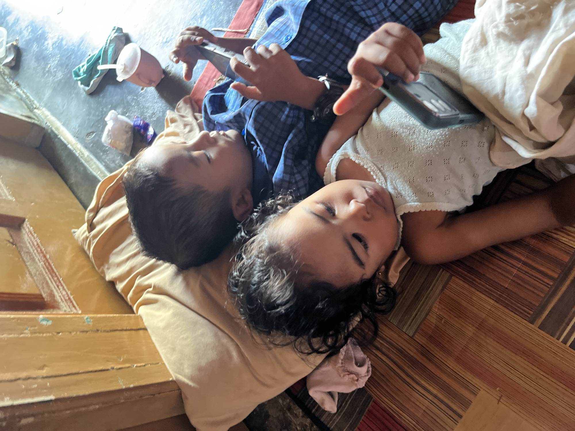 Two young children lie on their backs on a floor. The child on the left, wearing a blue plaid shirt, has their head resting on a tan pillow. Their eyes are closed, and they are holding a smartphone with both hands. The child on the right, wearing a white textured top, has their head also on a tan pillow. Their eyes are closed, and their head is turned slightly away from the viewer. A light-colored blanket or cloth is draped over the child on the right. In the background, on a blue surface, are various items including a plastic cup with a lid, a green cloth, and a small plastic bag. The floor has a striped pattern. The environment appears to be indoors.