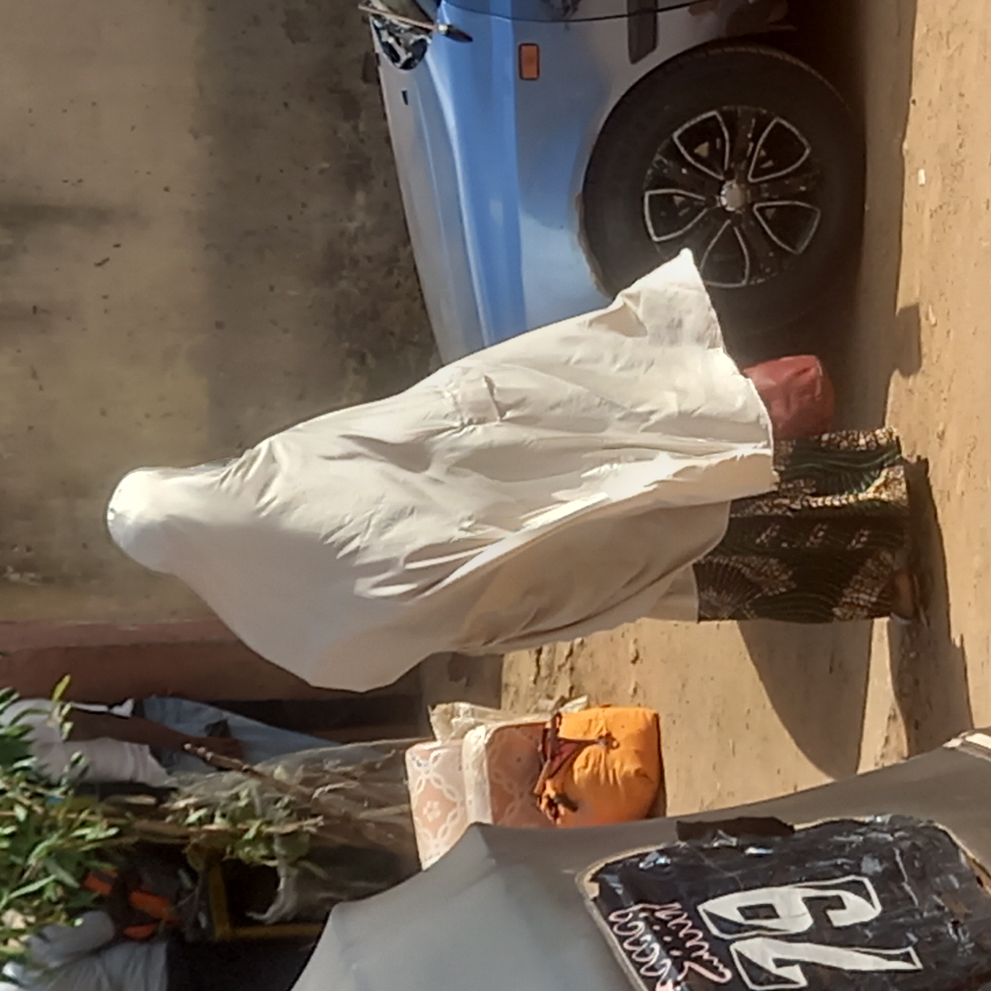 The image appears to show an individual covered in a white cloth, possibly a burial shroud, near a silver-colored vehicle. They are wearing a patterned skirt. The scene takes place outdoors on a dirt surface next to a car, and various items are also present, including plants, cushions, and a covered object with the number "76" on it. Based on the prompt information, it is likely the location is Sardauna, Nigeria.