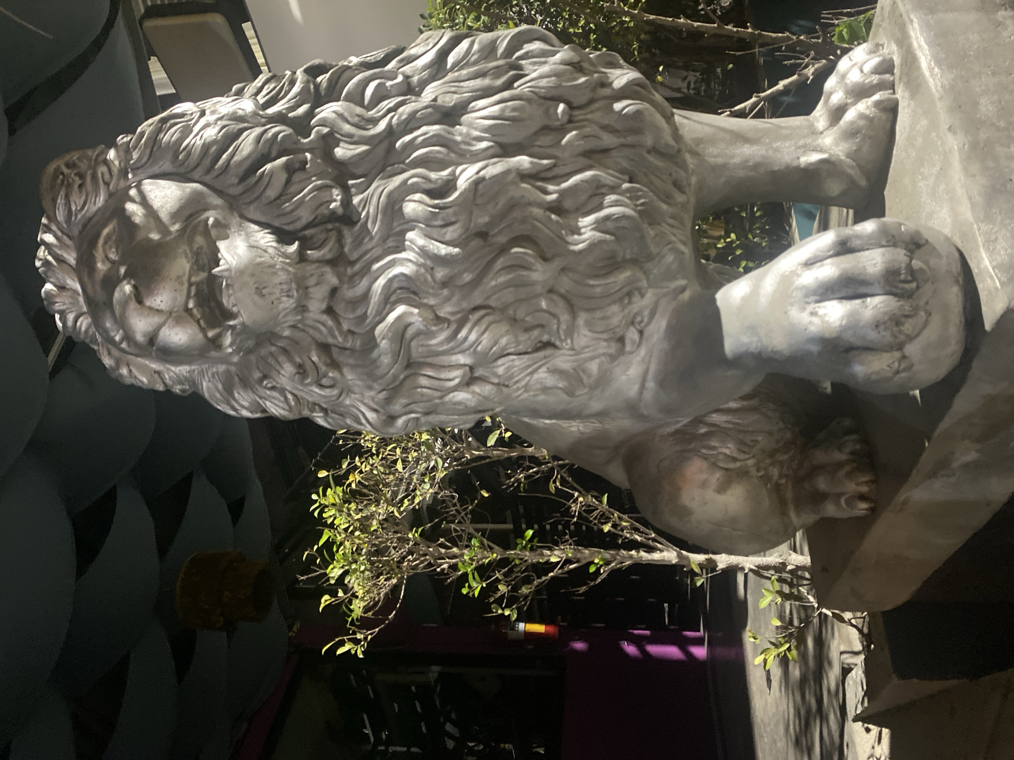 A silver-colored lion statue stands on a plinth. The statue is highly detailed, showing the lion's mane and facial features clearly. The statue is positioned outside; there is a small tree with green leaves behind the lion, and parts of a building can be seen in the background. A portion of a chair and decorative blue objects are visible. A purple wall is also apparent behind the tree. The location is Baan Don Sai, Thailand, as evidenced by the metadata. The scene appears to be at night or in a very dimly lit environment.