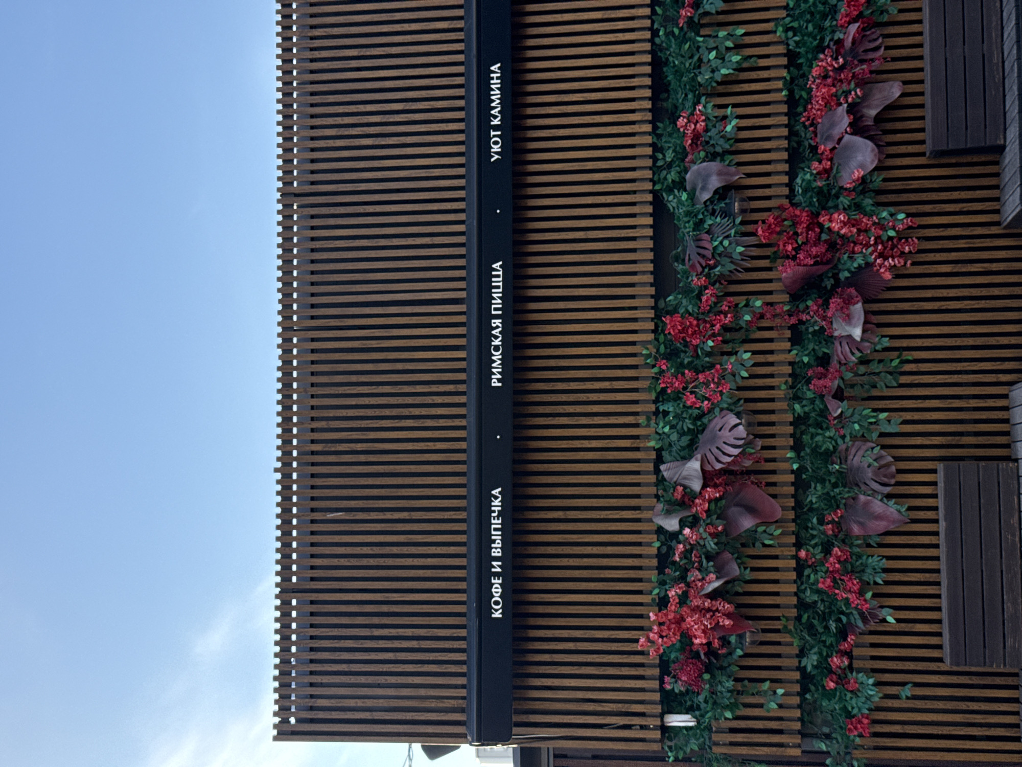 The image, originally captured in portrait orientation and rotated 90 degrees clockwise, depicts the exterior facade of a building under a bright, clear blue sky. The building's surface is composed of vertical wooden slats, creating a distinct, textured pattern.
A prominent black sign, integrated vertically into the slatted design, features white Cyrillic text. From top to bottom (which would be left to right if the image was upright), the text reads: "УЮТ КАМИНА" (meaning "Cozy Fireplace" or "Fireplace Comfort"), "РИМСКАЯ ПИЦЦА" (meaning "Roman Pizza"), and "КОФЕ И ВЫПЕЧКА" (meaning "Coffee and Pastries"). This signage indicates the establishment is likely a cafe or restaurant specializing in coffee, pastries, and Roman-style pizza, possibly with a cozy theme.
The facade is richly adorned with artificial decorative elements, including extensive green foliage and clusters of vibrant red and burgundy flowers and leaves. These decorations are arranged in two prominent vertical strips, enhancing the building's aesthetic appeal. Large, broad leaves in deep red or purple tones are interspersed among the greenery. To the right of the floral arrangements, a dark, rectangular section of the building, possibly a window or a solid panel, is visible, continuing the overall contemporary design.
The bright blue sky suggests it is daytime with clear weather. No people or specific activities are visible in this exterior shot. The combination of modern architectural elements, inviting signage, and lush artificial greenery presents a welcoming facade for this establishment in Zelenogorsk, Russia.
