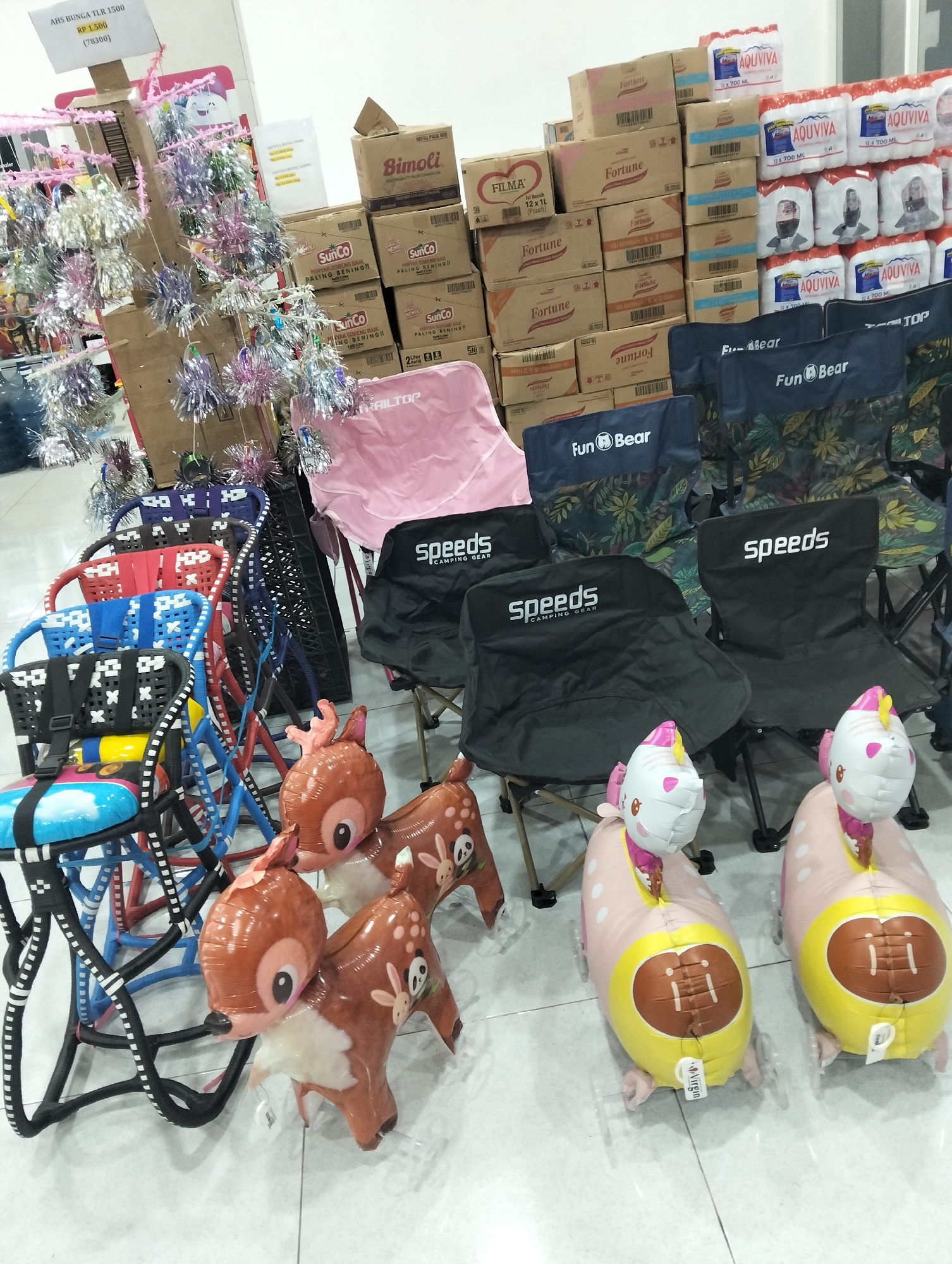 The image shows an indoor retail setting, likely a store in Kedungrejo, Indonesia, displaying various products. In the foreground, there are several colorful children's chairs made of woven plastic and metal frames, stacked and arranged. Two large, inflatable balloons in the shape of deer and two in the shape of characters with animal-like features and football-shaped lower bodies are also prominent.
Behind the chairs and balloons, there are several black folding camping chairs with the brand "Speeds" and "Fun Bear" printed on them. Further back, stacks of cardboard boxes are visible, some branded with "Fortune," "SunCo," and "Filma." There are also packs of bottled beverages, likely water, branded "AQUVIVA." In the upper left corner, a price tag is visible, reading "AHS BUNGA TLR 1500 (78300)." The environment appears to be a well-lit retail space with tiled flooring. No people are present in the image.