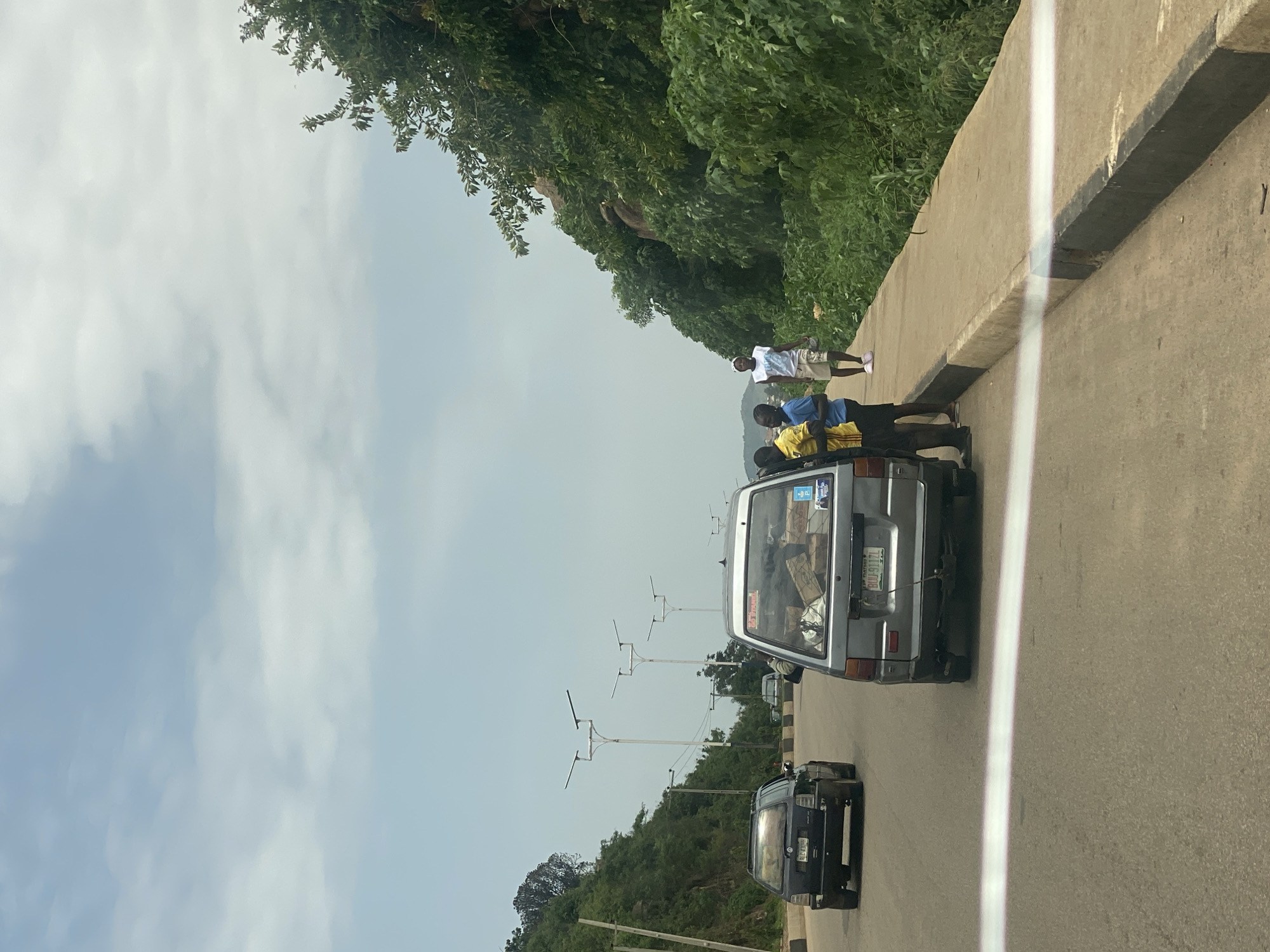 The image depicts a roadside scene in Gura Top, Nigeria. The orientation of the image is such that the sky and landscape are on the left, and the road is on the right.
In the foreground, a silver van is visible, loaded with boxes which are strapped to the roof and visible through the rear window. The van's license plate reads "BUD 9170". Following behind the silver van is a black van, its license plate obscured by glare.
On the left side of the road, a concrete barrier separates the road from a steep, tree-covered embankment. Several people are standing on the road near the silver van. One person, wearing a white t-shirt and shorts, stands on the concrete barrier. Below him, closer to the van, are three individuals, two of whom appear to be young boys. They are standing on or near the van, possibly loading or unloading items.
In the background, several streetlights with a distinctive design are visible along the side of the road. The sky is overcast with scattered clouds. The overall scene suggests a daytime setting, possibly with mild weather. The presence of multiple people and a loaded van might indicate transport of goods or people.