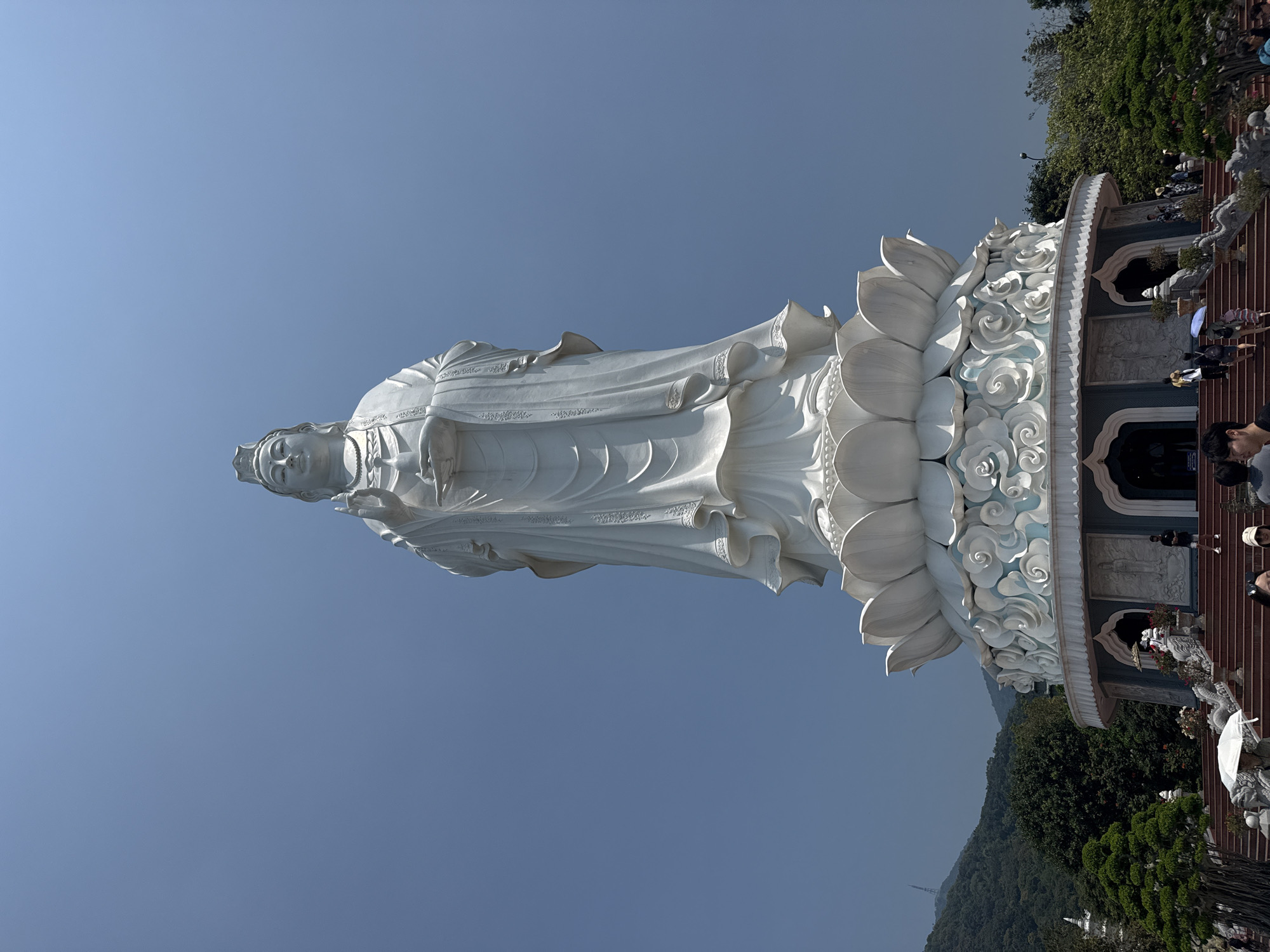 The image depicts the towering white statue of Guanyin (Lady Buddha) located at Linh Ung Pagoda on Son Tra Peninsula, Đà Nẵng, Vietnam. The statue, rendered in white stone or concrete, stands prominently against a clear, vibrant blue sky. It is depicted in a standing posture, with its right hand raised in a gesture of blessing and its left hand holding a small vase. The deity wears an elaborate crown, a necklace, and flowing robes with intricate relief details.
The statue rests on a multi-tiered pedestal, featuring a large white lotus flower base atop a circular structure adorned with white cloud-like motifs and smaller relief carvings. At the ground level of the pedestal, several arched entrances are visible. A wide, multi-tiered staircase, reddish-brown in color, leads up to the base of the statue.
Approximately two dozen individuals, appearing to be visitors or tourists, are visible on these steps and around the statue's base. They are dressed in casual attire, with some holding white umbrellas for sun protection as they ascend and descend the stairs or gather.
To the left and in the background, densely forested green mountains rise, with a tall, slender communication or observation tower visible on a distant peak. Another smaller white structure, possibly a stupa, is partially visible among the trees on the left. Green foliage, including sculpted bonsai-style trees, flanks the staircase leading to the statue. The scene is illuminated by bright daylight under a clear sky, indicating fair weather conditions.