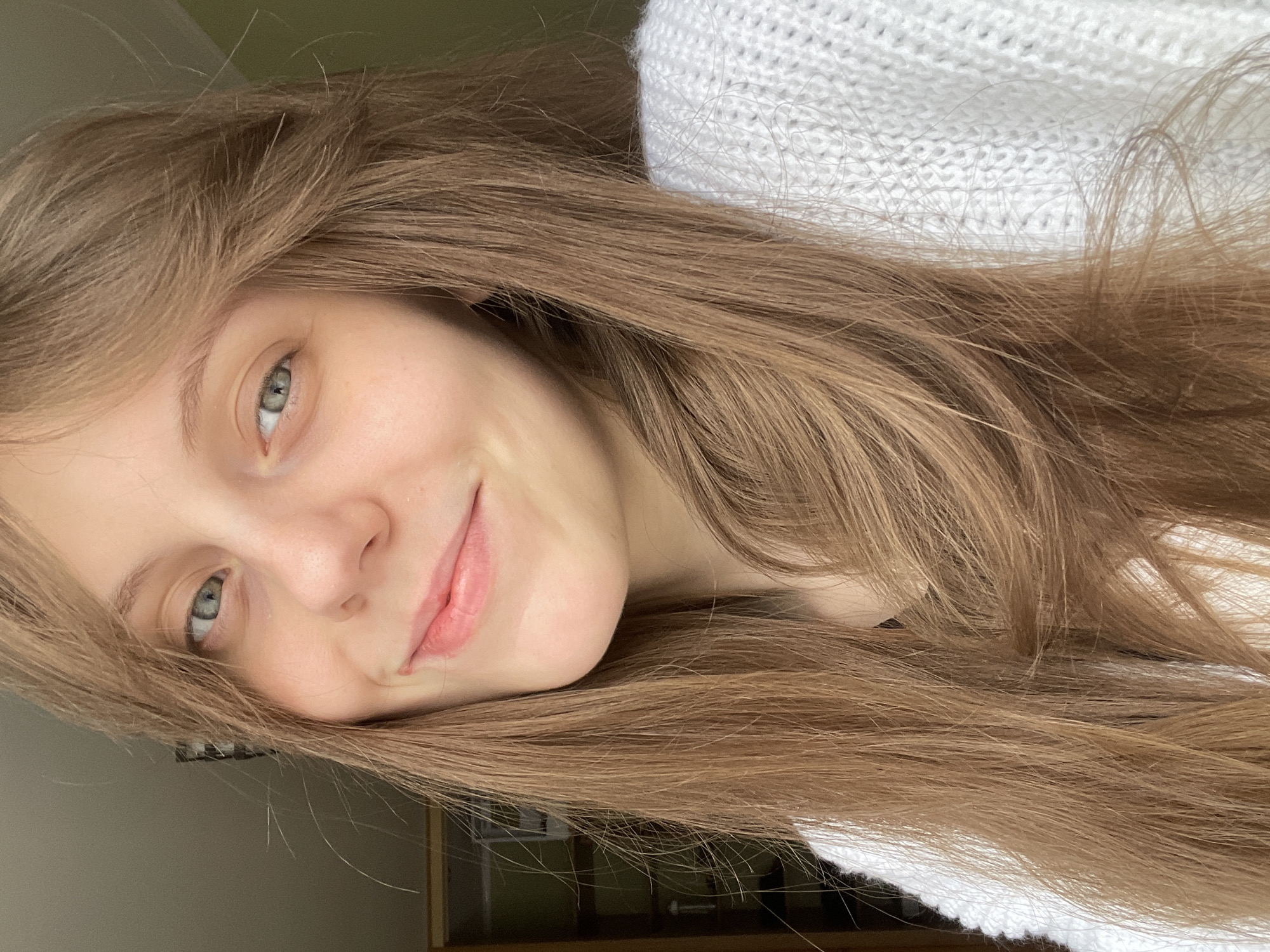 The image is a close-up, eye-level selfie of a young woman with long, light brown hair. She is looking directly at the camera with a gentle smile. Her eyes are a light blue-green color. She is wearing a white knitted sweater. The background is slightly blurred, showing what appears to be an indoor setting with a shelf unit on the left and a light green wall on the right. There is no discernible information about the location being in Ufa, Russia, or any specific urban context. The scene is personal and domestic.