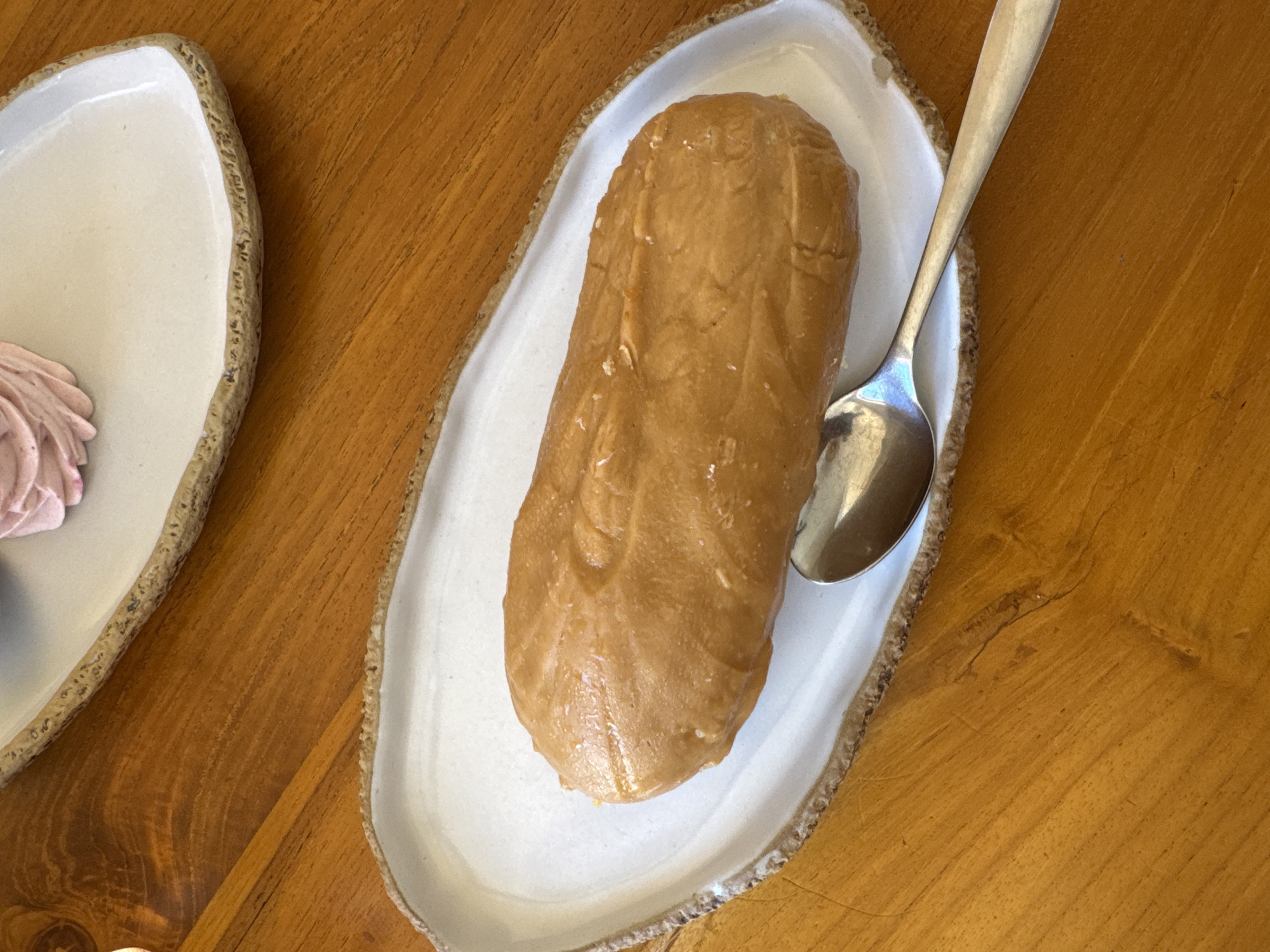 A tan-colored loaf of dessert, possibly peanut butter flavored, is served on an oblong off-white plate with a speckled, tan-colored rim. A silver spoon rests on the right side of the plate, partially covering the dessert. Another plate containing a pink-colored dessert is partially visible to the left of the main plate. The scene takes place on a light brown wooden table. The location is Kuta Selatan, Indonesia.