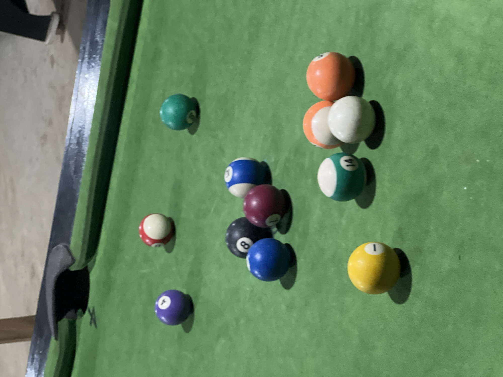 The image displays a billiards or pool table in an indoor setting, likely a game room or leisure space in Minna, Nigeria. The table has a vibrant green felt surface, showing some minor wear and specks, with dark rails and a prominent black corner pocket visible on the left.
Several pool balls are scattered across the felt, indicating that a game has either just begun with a break shot or is currently in play. The lighting appears to be artificial, casting distinct, elongated shadows from each ball, suggesting a light source positioned above and to the right of the frame.
A total of twelve pool balls are visible, including:
* A solid yellow ball with the number '1' (bottom right)
* A solid blue ball with the number '2' (mid-left)
* A red and white striped ball with the number '3' (mid-left, near the pocket)
* A solid purple ball with the number '4' (bottom-left)
* A solid orange ball with the number '5' (top-right cluster)
* A solid green ball with the number '6' (top-left)
* A solid maroon ball with the number '7' (center)
* A solid black ball with the number '8' (center, below the maroon '7')
* An orange and white striped ball with the number '9' (top-right cluster)
* A blue and white striped ball with the number '10' (center, right of the '8' ball)
* A green and white striped ball with the number '14' (top-right cluster, below the cue ball)
* A plain white cue ball (top-right cluster, below the '9' ball).
No people or discernible emotions are visible, the focus being solely on the game setup.