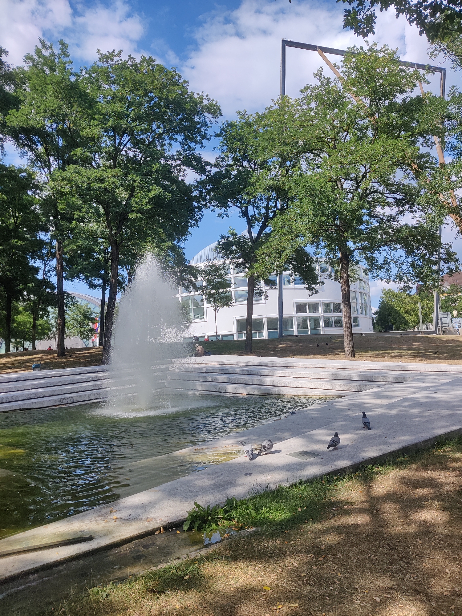 The image shows a modern architectural park in Bielefeld, Germany, featuring a water fountain as the central element. The fountain is located within a multi-tiered concrete structure that borders a body of water. Several pigeons are scattered on the concrete walkway and near the water's edge. In the background, a modern white building with large windows is visible, surrounded by lush green trees. The sky above is blue with scattered clouds. The overall scene depicts a tranquil urban outdoor space.