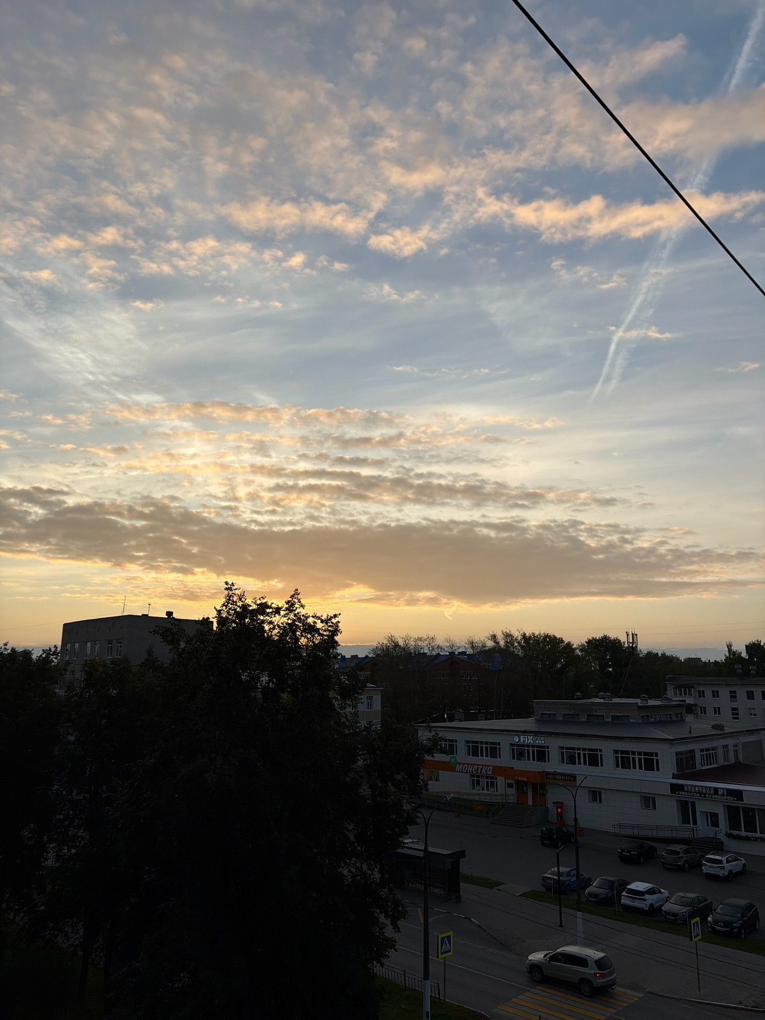 The image shows a view of a city street in Chaikovsky, Russia, at dusk. The sky is filled with scattered clouds illuminated by the setting sun, casting a warm, orange glow. A dark silhouette of trees is visible in the foreground, partially obscuring the view of buildings and a street below. Several cars are parked along the street, and a silver SUV is driving across a crosswalk. The buildings appear to be commercial establishments, with signage that is partially visible. The overall scene is calm and quiet, characteristic of a late evening in a residential or commercial district.