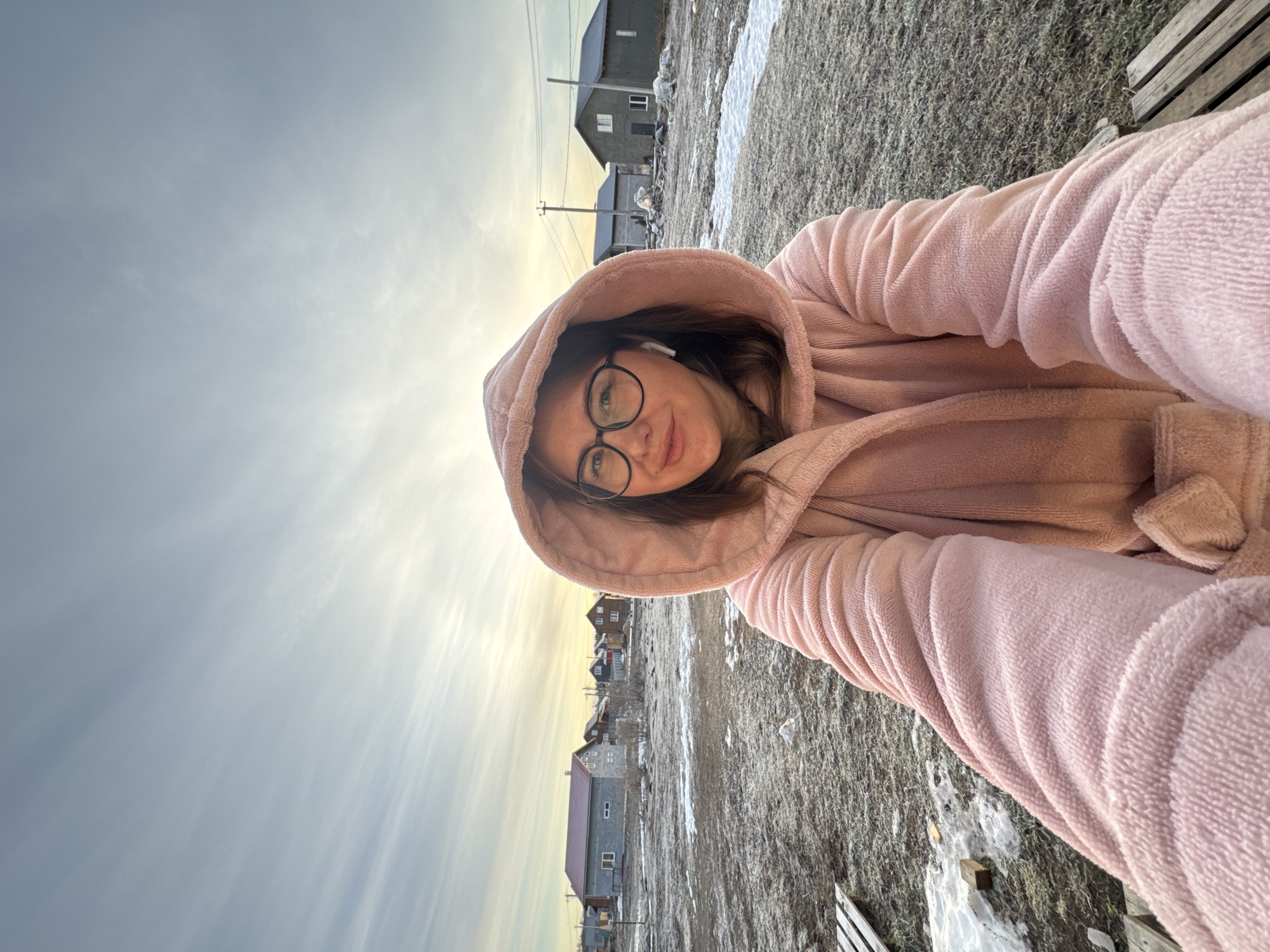 A woman wearing a pink hooded robe and glasses is taking a selfie outdoors in Ферма, Russia. There are several buildings, patches of snow, grass, and power lines visible in the background. The sky is overcast.
