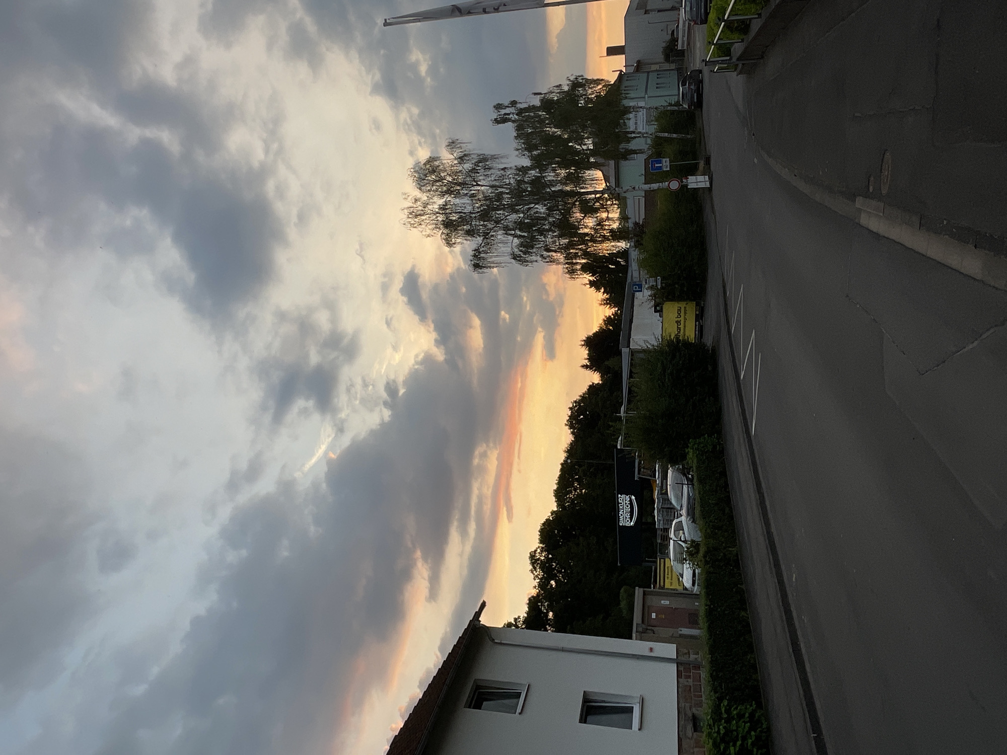 The image captures a view of a street in Neukirchen, Germany, during what appears to be sunset. The sky is filled with dramatic clouds, painted with hues of gray, white, and a warm orange-pink from the setting sun. A tall, slender tree with delicate branches stands prominently against the colorful sky.
In the foreground, the roof and upper story of a white house are visible, with two rectangular windows. A paved road curves to the right, bordered by a curb and a grassy verge. Along the roadside, various objects are present, including parked cars, a fence, and dense green bushes.
Further down the road, buildings and more trees line the street. A large, dark sign with white text is visible, which reads "SIMON KROGER BRÜCKENBAU." Another smaller yellow sign with text that appears to be "herdt bau" is also present. Traffic signs, including a blue sign with white lettering and a red and white circular sign, are also discernible. The overall atmosphere is one of a quiet evening in a suburban or semi-rural setting, with the striking cloud formation adding a touch of natural drama to the scene.