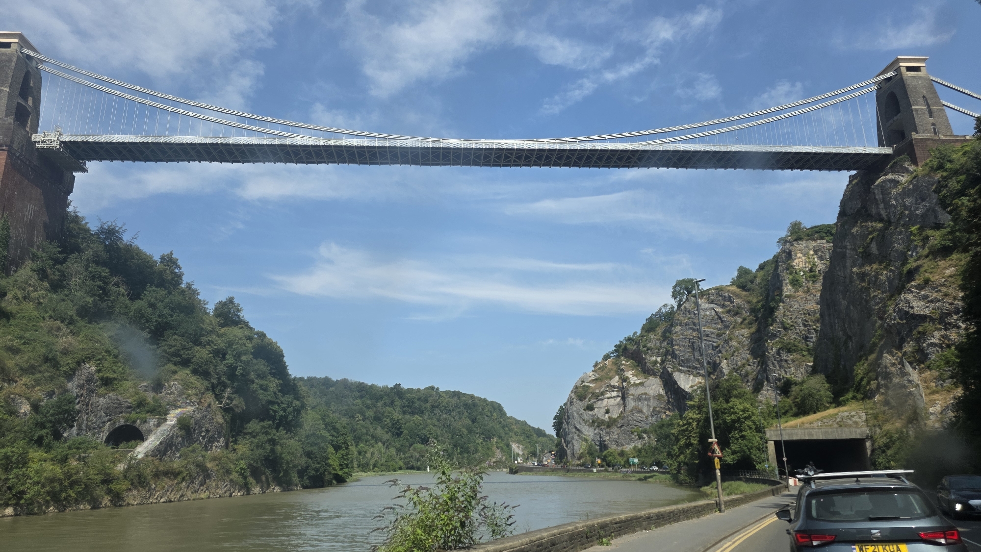 This is a medium shot, eye-level, outdoor, daylight, wide-angle shot of the Clifton Suspension Bridge in Bristol, United Kingdom. The bridge spans a river, with steep, rocky cliffs on either side covered in lush green trees. The sky above is a bright blue with scattered white clouds. In the foreground, a road runs alongside the river, with a dark grey car driving away from the camera. The car has a license plate that reads "WF21 KUA". Further down the road, a tunnel entrance is visible carved into the cliff face. The river itself is a calm, murky green. The overall scene depicts a majestic natural landscape enhanced by a famous man-made structure.