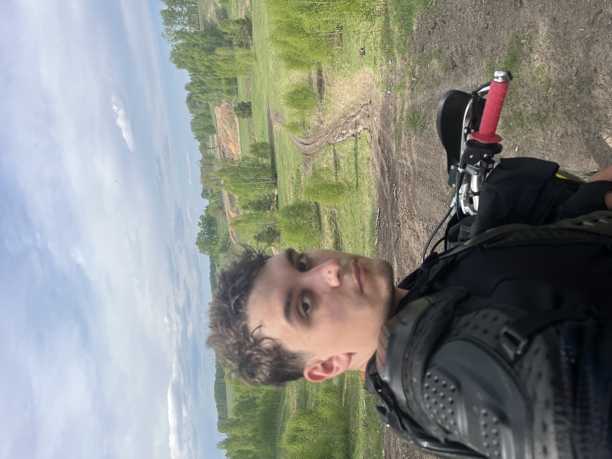 The image shows a person riding a motorcycle in Новочувашский, Russia. The individual is wearing protective gear. The background consists of a field and trees under a partly cloudy sky. A dirt path can be seen in the field. The motorcycle has red handlebar grips.