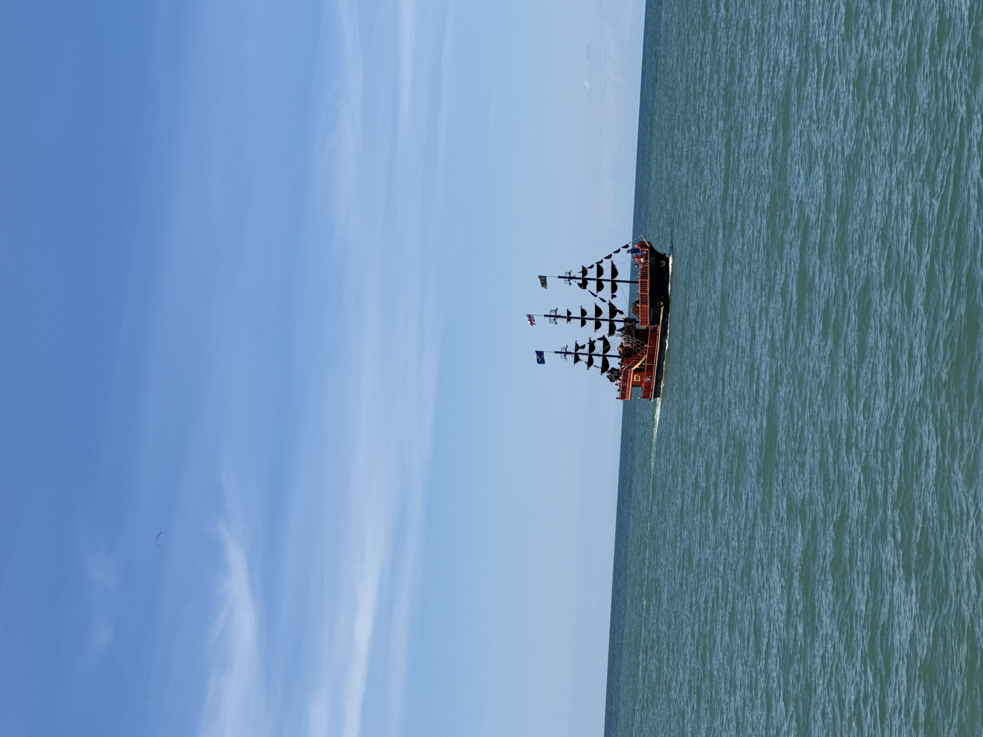 A sideways view captures a pirate ship sailing on the sea. The sky takes up most of the left side of the image, displaying a mix of blue shades with a few wispy clouds. The sea fills the right side of the image and is a greenish-blue color. The pirate ship is positioned on the horizon and has black masts and sails. Flags are visible on the top of the masts. The ship also has red and orange accents.