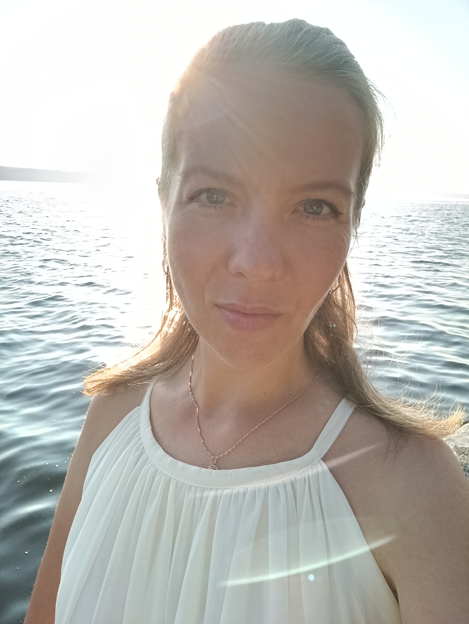 A woman is taking a selfie outdoors by the water. She has blonde hair pulled back and is wearing a white dress. The sun is bright, creating a glare in the image. The water appears to be a river or lake, with ripples on the surface and a faint shoreline visible in the distance. The location is the Kama River embankment in Chaikovsky, Russia, as indicated by the presence of the Kama River and the characteristic landscape.