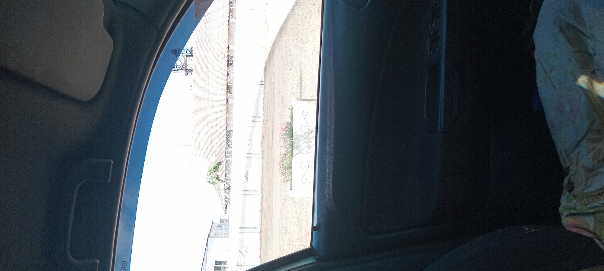 The image shows a view from inside a car looking out through the open window. The exterior scene features a white wall with a decorative pattern and barbed wire along the top, a white building with windows in the background, a white corrugated roof structure behind the wall, and some industrial equipment visible above the roofline. In the foreground of the exterior, there is a rectangular white planter box containing pink flowers. The interior of the car shows a dark-colored car door panel with window control buttons. A fabric-covered object is visible on the car seat in the lower portion of the image. The sky is bright and appears to be daytime. The location is specified as Koviahy, Ukraine.