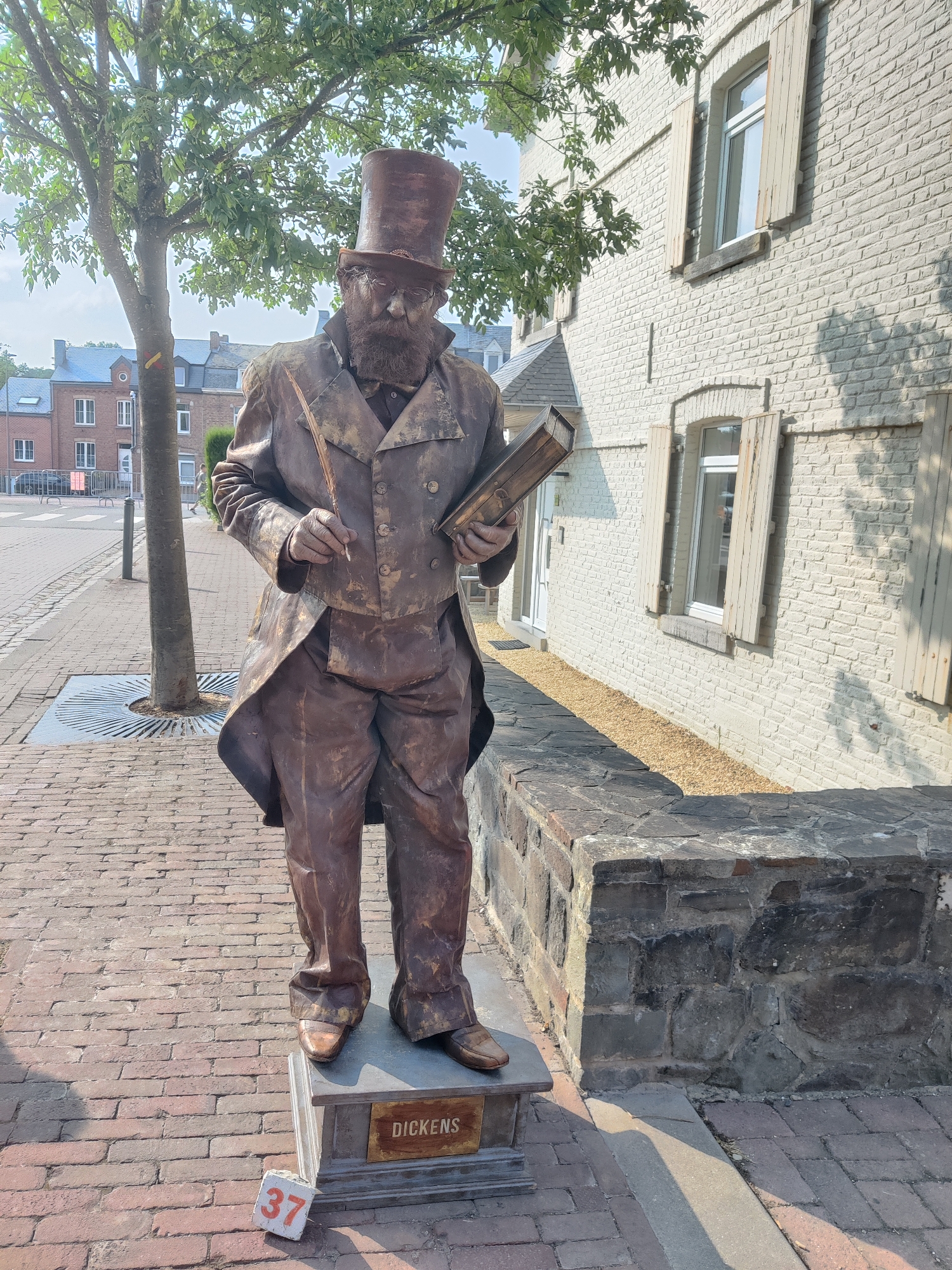 A statue of a man dressed in Victorian-era attire, complete with a top hat, stands on a brick sidewalk. The statue is bronzed, giving it a metallic appearance, and it holds a quill pen in its right hand and a book in its left. The statue is mounted on a pedestal with the name "DICKENS" inscribed on it. In front of the pedestal, a small concrete block with the number "37" painted in orange is placed on the ground.
The statue is positioned in front of a light-colored brick building with closed wooden shutters on the windows. A tree with green leaves stands to the left of the statue, casting a shadow on the sidewalk. The sidewalk is paved with bricks and a metal grate is visible around the base of the tree. In the background, there are more brick buildings, a street, and a few cars. A portion of a stone wall is also visible to the right of the statue.
The setting is an urban environment, likely a public space or a street in a town. The presence of the "DICKENS" inscription and the Victorian costume suggest that the statue may be related to Charles Dickens or a literary-themed attraction. The scene is illuminated by natural daylight, indicating it is daytime.
Based on contextual information, this image was taken in the city of Marche-en-Famenne, Belgium, specifically on the Rue Jean-Jaurès.
