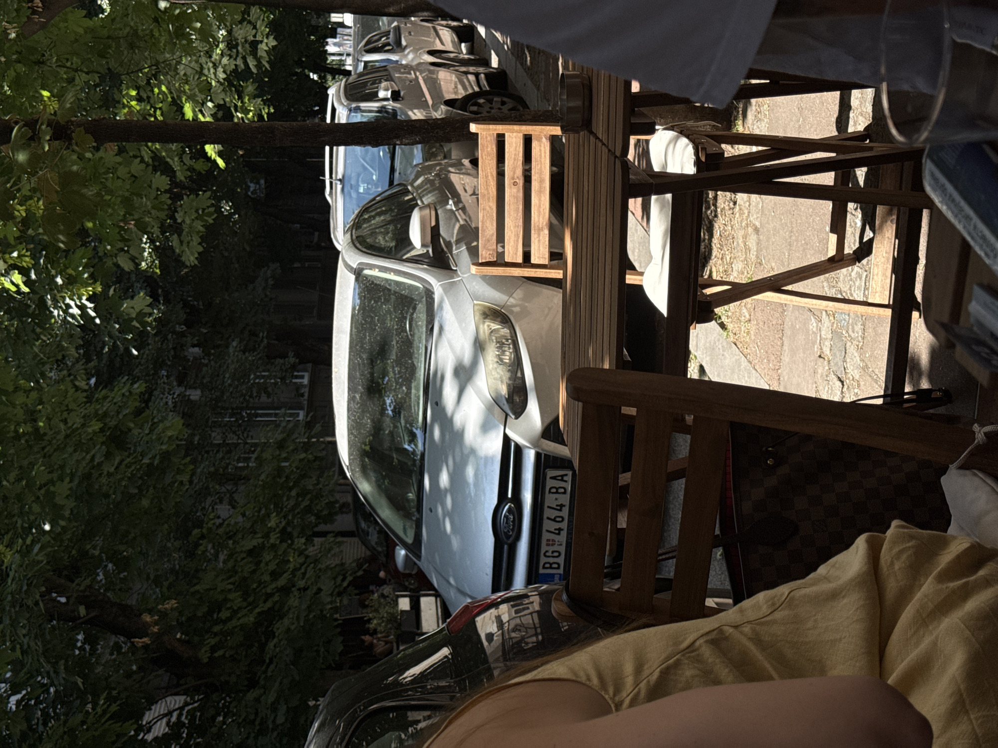 The image depicts a street scene in Belgrade, Serbia, likely in a cafe setting. In the foreground, a wooden table and chairs are visible, with a checkered designer bag placed on one of the chairs. A person wearing a yellow dress is seated on another chair, partially obscuring the view. The background shows several parked cars on the street, including a silver Ford Focus with the license plate "BG 464-BA". Trees with dense foliage line the street, casting shadows on the cars and the cafe furniture. The overall impression is that of a casual outdoor dining or waiting area on a sunny day in the city.