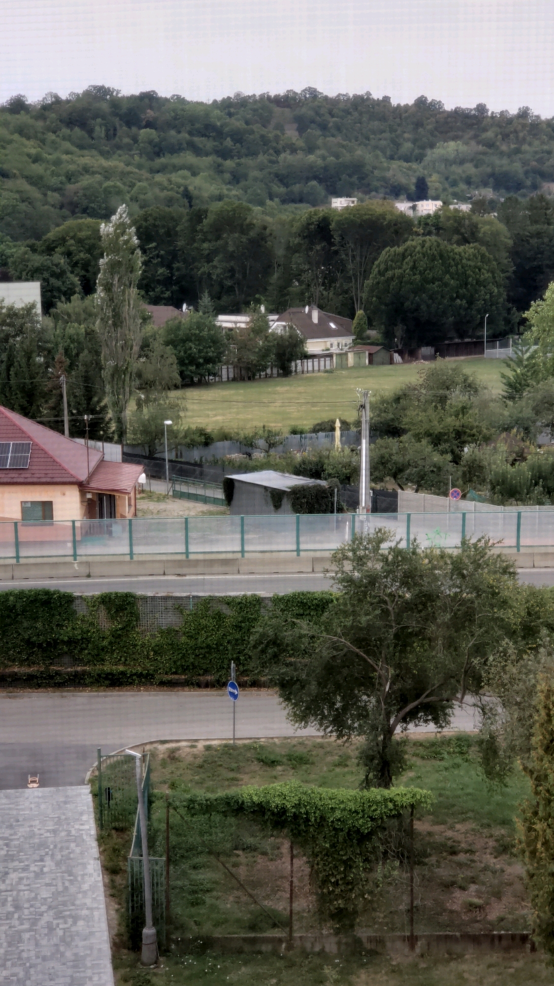 The image shows a residential area on a hill in Prešov, Slovakia, possibly overlooking a highway or major road. In the foreground, a grey paved pathway leads to a low-angle shot of a house with a red roof and solar panels. Further back, a highway barrier is visible, with a street and a traffic sign on it. Beyond the road, there are more houses and a large expanse of green grass. The background is dominated by a lush green forest-covered hill. The sky is overcast.