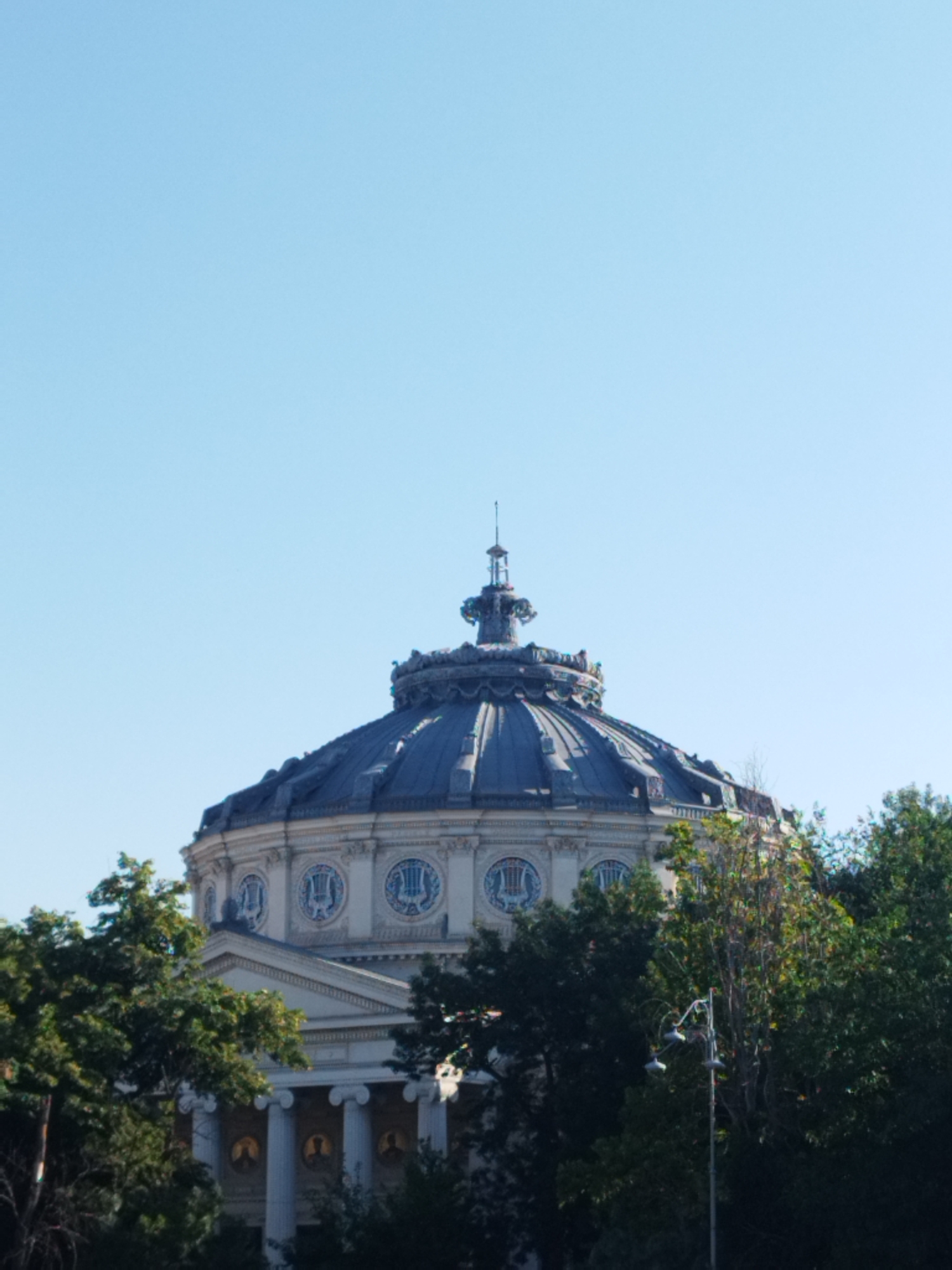 The image shows the Romanian Athenaeum in Bucharest, Romania, a grand neoclassical concert hall. The building's distinctive dome and facade are visible, bathed in natural light under a clear blue sky. Lush green trees surround the base of the building, partially obscuring its lower levels and adding a natural element to the urban landscape. A street lamp with multiple lights stands in front of the trees, adding a modern touch to the scene. The photograph is taken from a low angle, emphasizing the architectural grandeur of the Athenaeum.