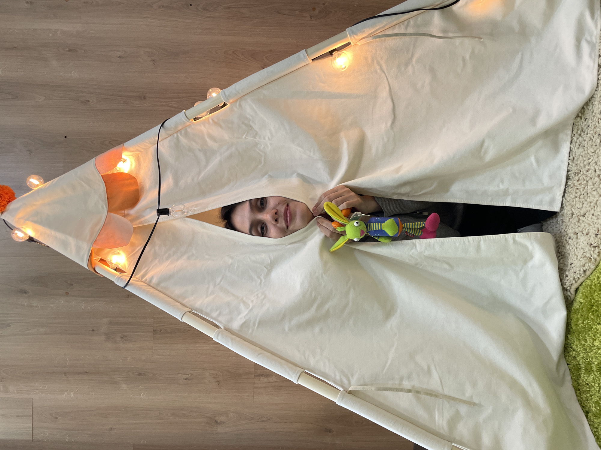 A woman lies inside a teepee tent, holding a toy. The teepee is lit with string lights. The flooring on the left is wood-patterned. On the right, there is a textured rug. The location is not specified in the image.