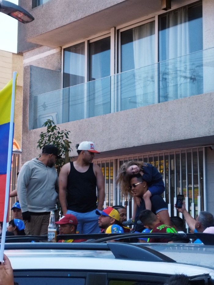 The image depicts an urban outdoor scene in Santiago, Chile, capturing a public gathering or procession during daylight hours. A modern, multi-story building with a light-colored facade, featuring large windows and a clear glass balcony railing on an upper floor, serves as the background. A street lamp extends from the upper left.
In the foreground, the white roof of a vehicle is visible, with several individuals standing through what appears to be a sunroof or open top. A hand on the far left holds a flag pole supporting a flag with vertical stripes of yellow, blue, and red. Among the individuals in the vehicle, one man in a red baseball cap holds a light-colored beer bottle. Next to him, another man wears a segmented cap in yellow, blue, and red colors. Further right, a man wearing sunglasses and a dark polo shirt carries a young child with long blonde hair on his shoulders; the child is leaning forward. Behind them, a man holds a black smartphone vertically, seemingly recording.
In the midground, to the left of the vehicle, two men stand on a sidewalk. One wears a light grey hooded sweatshirt and a dark cap, with dark gloves. The other wears a black tank top, blue shorts, and a white baseball cap with red lettering ("BOS"). A small leafy plant is situated between these standing men and the vehicle. People are looking in various directions, indicating active participation or observation of an event.