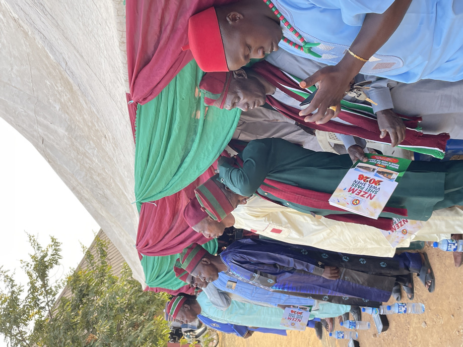 A row of seated men wearing various traditional Igbo attire including red and green striped hats, colorful gowns, and beaded necklaces, are under a tent draped in maroon and green fabric. One man in the row is holding a brochure titled "NZEM 2028". Several water bottles are placed on the ground near their feet. A green tree and clear blue sky are visible in the background, suggesting an outdoor event.