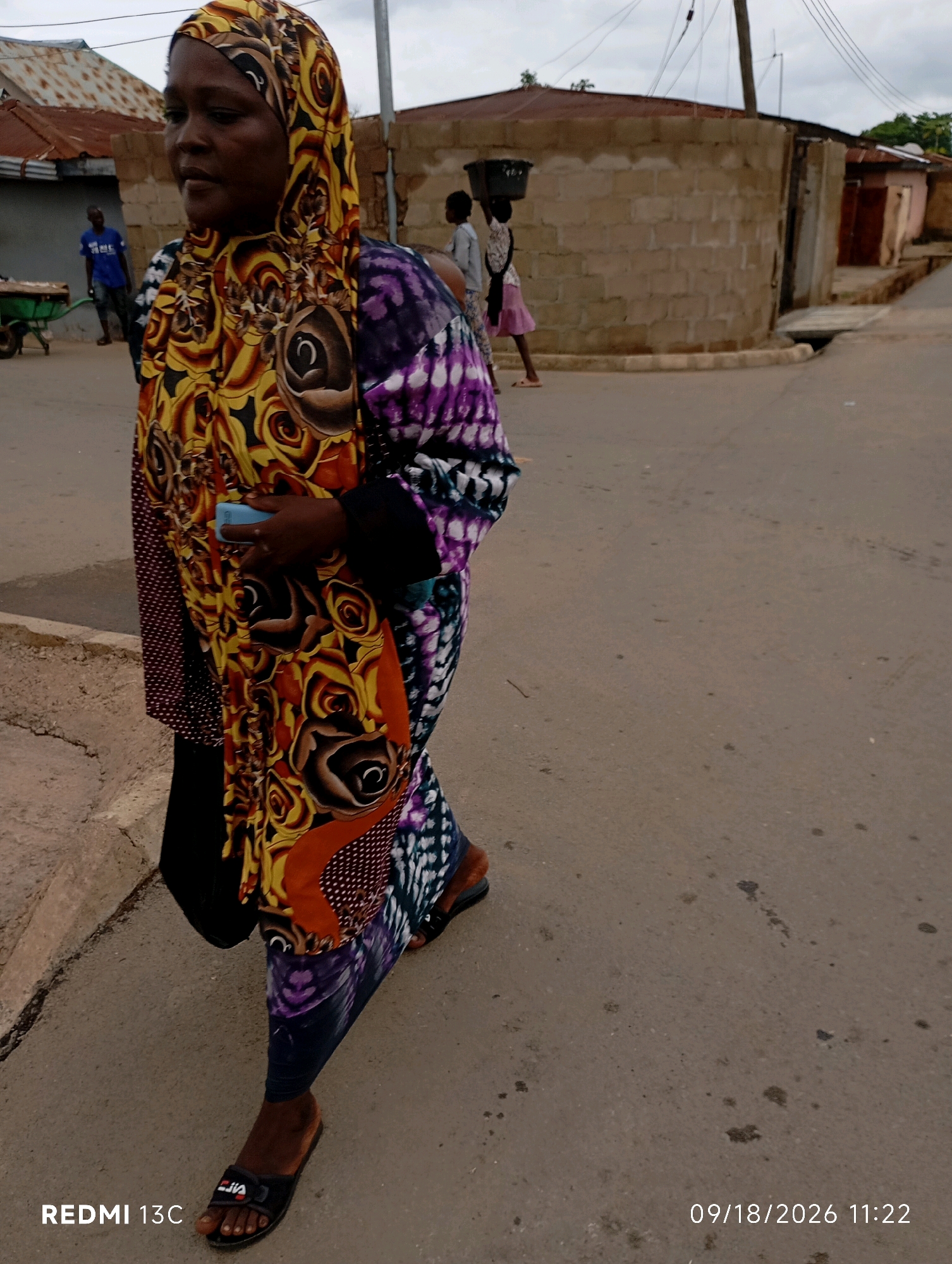 In the city of Shendam, Nigeria, a woman walks down a dusty street, her vibrant, patterned attire a striking contrast to the muted tones of the urban surroundings. She wears a headscarf and a long, flowing dress, adorned with a colorful print of roses and tie-dye patterns. Her right hand clutches a light blue mobile phone, as if taking a break from a call or checking notifications. Her feet are clad in black flip-flops with a white logo. Behind her, another young woman walks with a black bucket balanced on her head, while in the distance, a man in a blue t-shirt stands near a green wheelbarrow. The street is lined with brick buildings and corrugated iron roofs, hinting at the modest architecture of the area. The sky above is overcast, suggesting a cloudy day. The image captures a moment of everyday life in Shendam, a snapshot of human activity against the backdrop of a typical Nigerian street scene.