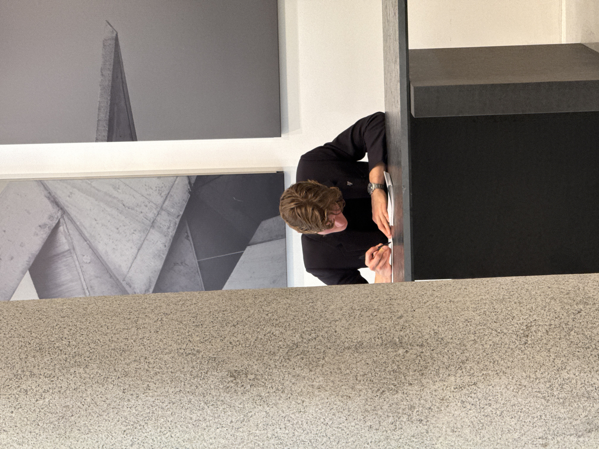 The image shows a man with blond hair, wearing a dark long-sleeved shirt and a watch on his left wrist. He is standing and appears to be writing on or interacting with a dark, rectangular structure. This structure has a dark vertical support. Behind the man, there are two black and white images mounted on a wall. The images are geometric or architectural in nature. The lower portion of the frame shows a textured, light-colored surface, likely a wall or countertop. The location is suggested to be Milan, Italy, based on the style of architecture.