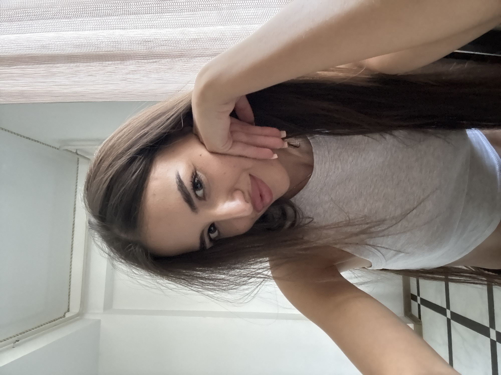 A young woman with long, dark brown hair is shown in a close-up selfie. She is wearing a light gray tank top and leaning her head on her hand. Her eyes are open and she is looking towards the camera with a slight smile. Her lips are slightly parted and appear to be colored with a light lipstick. Behind her, a sheer, light-colored curtain is visible on the right side of the frame, and a white wall with a ceiling molding is on the left. The floor in the lower left corner has a black and white checkered pattern. The overall setting appears to be indoors, possibly a room in a residential building. No specific location in Chișinău, Moldova can be determined from the image.
