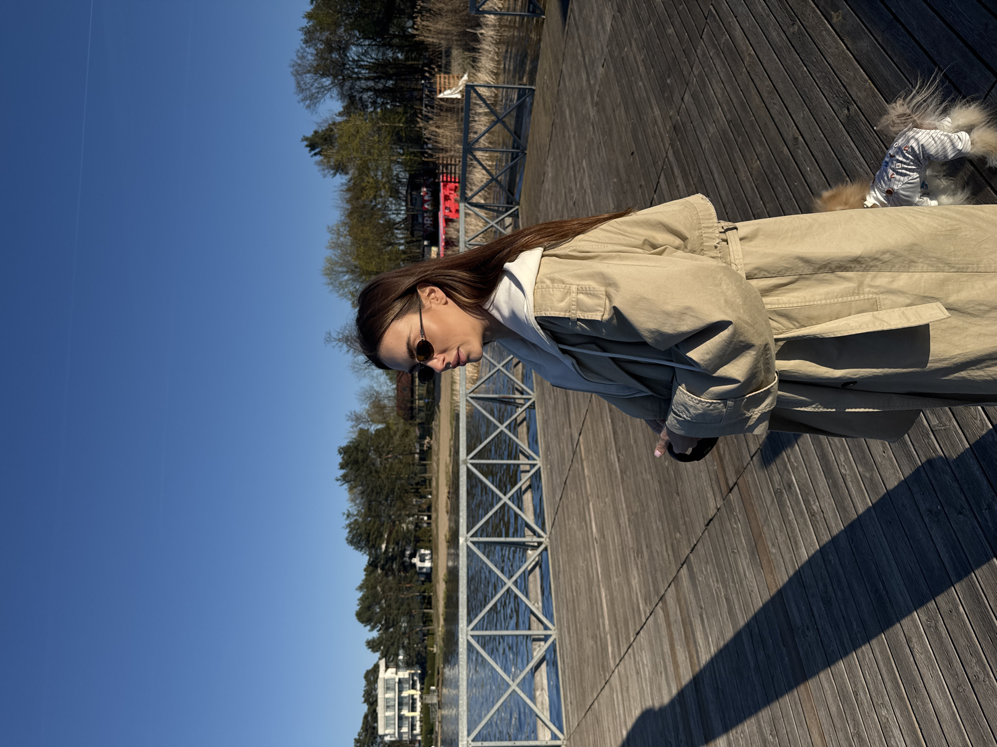 A woman in a beige trench coat and sunglasses stands on a wooden pier next to a small dog in a striped outfit. The pier extends over a body of water with buildings and trees visible on the opposite shore. A metal railing runs along the edge of the pier. The scene is located in Białobrzegi, Poland.