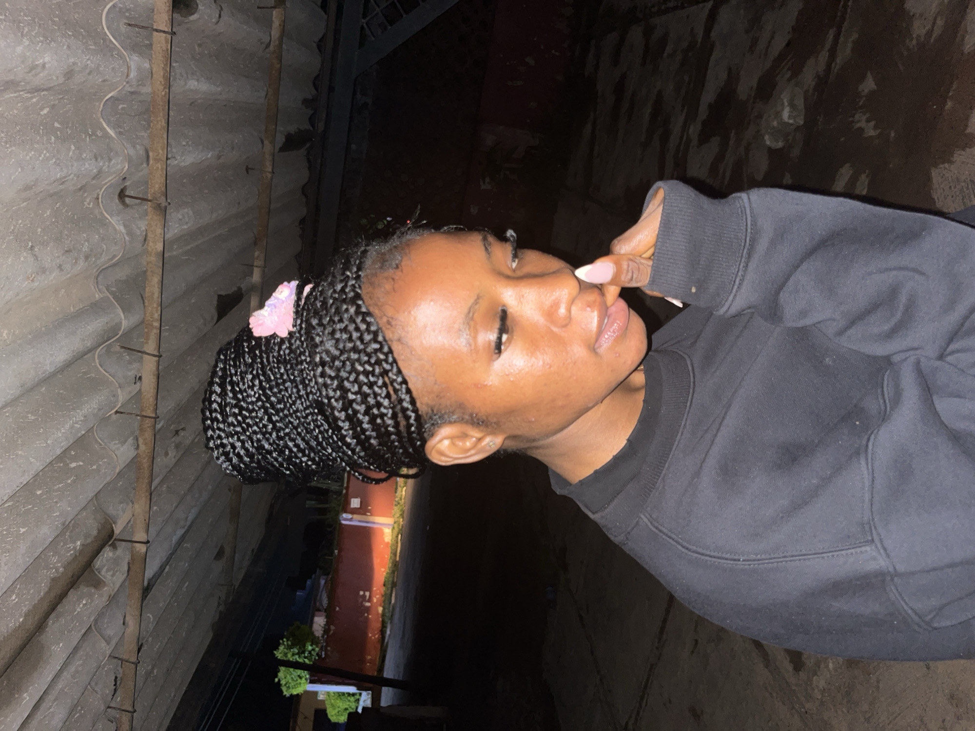 A young Black woman with braided hair styled in a bun, adorned with a pink hair accessory, is captured in a medium shot. She wears a dark crewneck sweatshirt. Her head is tilted downwards, and her right hand is raised to her mouth, with her fingers touching her lips. Her eyes are cast downwards. She is positioned outdoors, with a corrugated metal roof structure overhead. The background is dimly lit and appears to be an urban setting, possibly in the city of Jos, Nigeria, with hints of red-walled buildings and vegetation visible. The ground is rough and uneven.