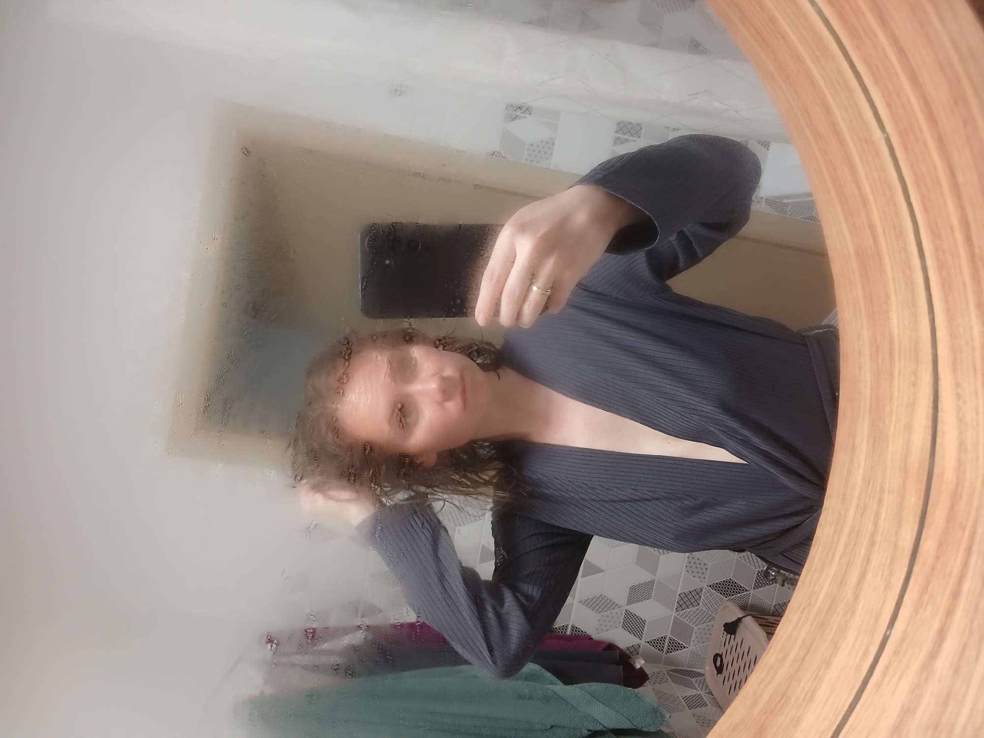The image is a portrait of a young woman in a bathroom, taken in Yekaterinburg, Russia. She is standing in front of a mirror that is partially fogged up, with water droplets visible on its surface. The woman is holding a smartphone in her right hand, taking a selfie. She is wearing a dark gray ribbed robe that is open at the neck, revealing her décolletage. Her hair is curly and appears to be damp, and she is gently touching it with her left hand. Her facial expression is relaxed and slightly playful.
In the background, reflected in the mirror, is a bathroom interior. The wall behind the woman is tiled with a geometric pattern of gray and white squares. There are some folded towels visible on a surface, in shades of teal, purple, and dark red. A light-colored wall is also visible, suggesting a room behind the mirror. The lighting in the image suggests it is daytime, and the fog on the mirror implies the presence of steam, likely from a shower. The overall mood is casual and intimate, capturing a moment of personal grooming. The image is oriented vertically, with the woman's head towards the top and her body extending downwards.