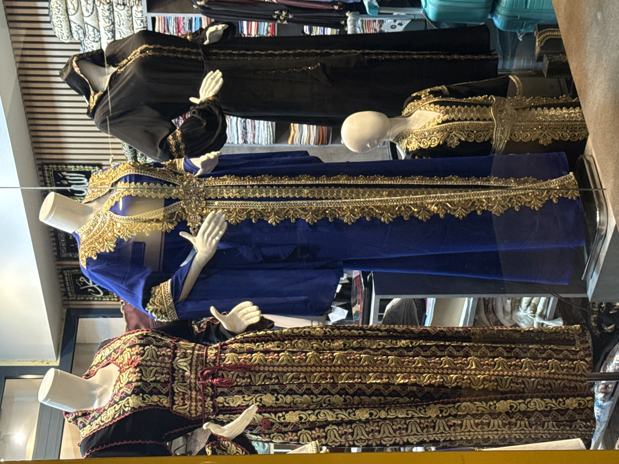 The image depicts a storefront window display of women's traditional clothing in Ilidža, Bosnia and Herzegovina. Three mannequins are prominently featured, showcasing elaborate garments. The mannequin on the far left wears a long, black kaftan with intricate gold and red embroidery that covers the entire dress, extending to the sleeves. A thin, decorative belt cinches the waist. The middle mannequin is dressed in a vibrant royal blue kaftan, also adorned with extensive gold embroidery along the neckline, sleeves, and down the front. A wide, ornate gold belt is worn over this kaftan. The mannequin on the far right is a covered figure in a black abaya with a hood, featuring gold trim on the edges and cuffs. Another smaller mannequin in the foreground, placed lower, wears a black garment with a gold embroidered chest piece and belt. Behind the mannequins, shelves are visible, stacked with folded textiles and what appear to be more garments, along with some decorative elements, suggesting a retail environment. The overall impression is of a shop selling richly decorated, culturally significant clothing.