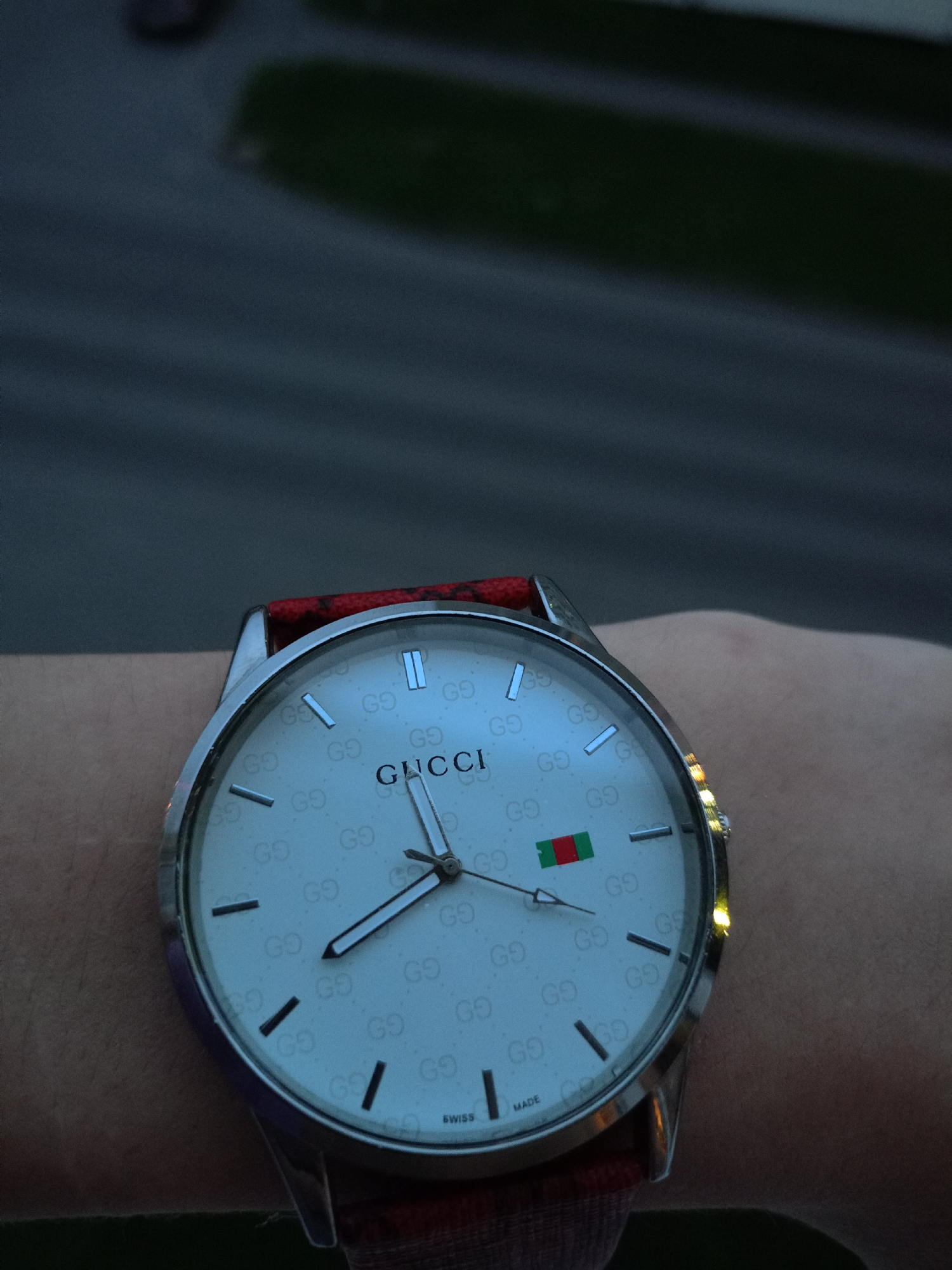 A close-up shot shows a Gucci watch with a white face and a red band being worn on a person's wrist. The watch face has the Gucci logo prominently displayed and a green and red stripe detail. The background is blurred and dark, suggesting an outdoor setting at dusk or night. The image does not contain any information that allows for the identification of the specific location in Petrozavodsk, Russia.
