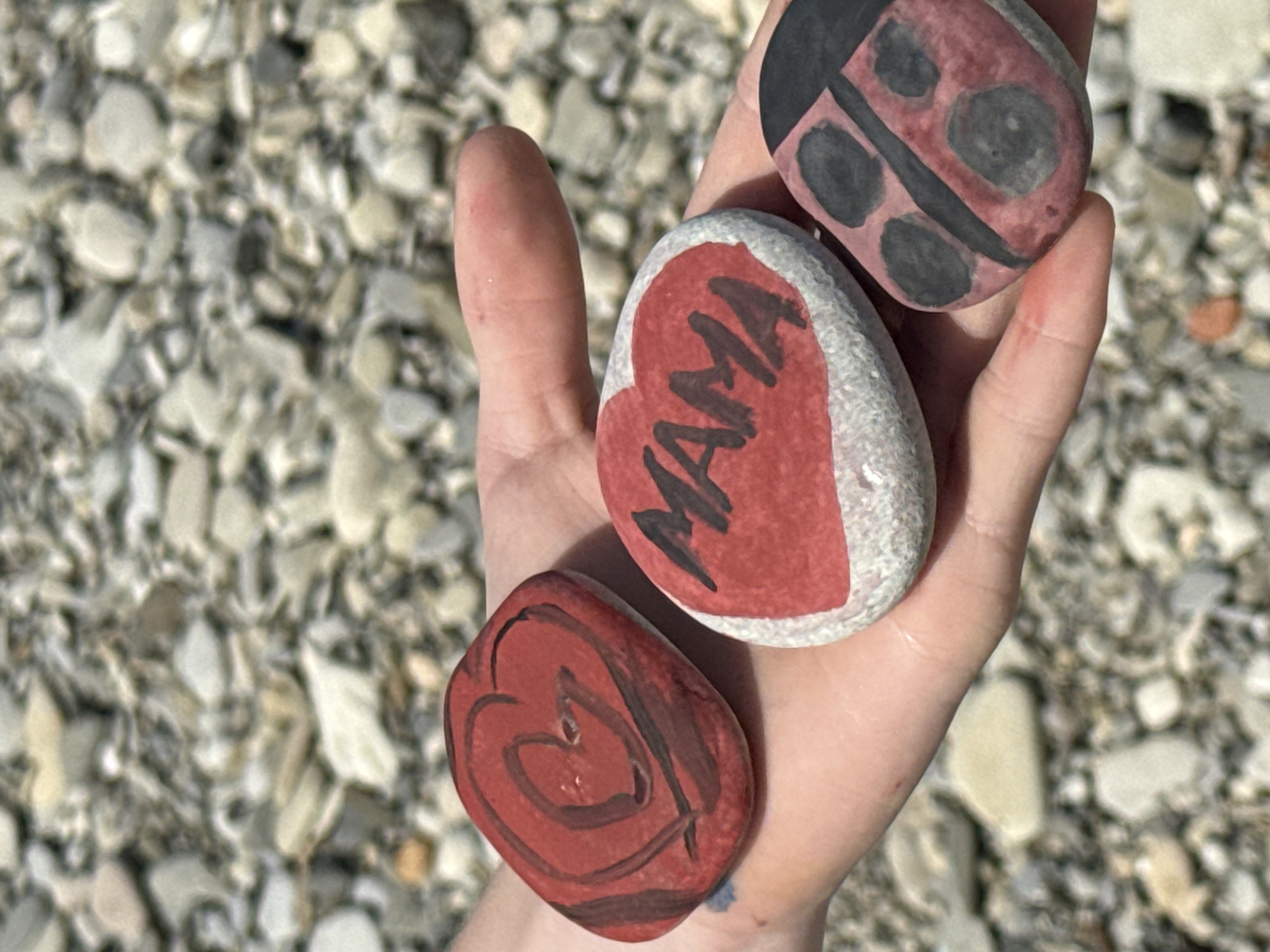 A light-skinned hand holds three painted stones in an outdoor setting in Nebug, Russia. The background is blurred, revealing numerous small, light-colored pebbles. The leftmost stone is reddish-brown with a darker red heart outline. The central stone is oval, grey with a red painted heart, bearing the word "MAMA" in black capital letters. The rightmost stone is reddish-pink, painted to depict a ladybug with a black head, a central black line, and four black spots. Natural daylight illuminates the scene.