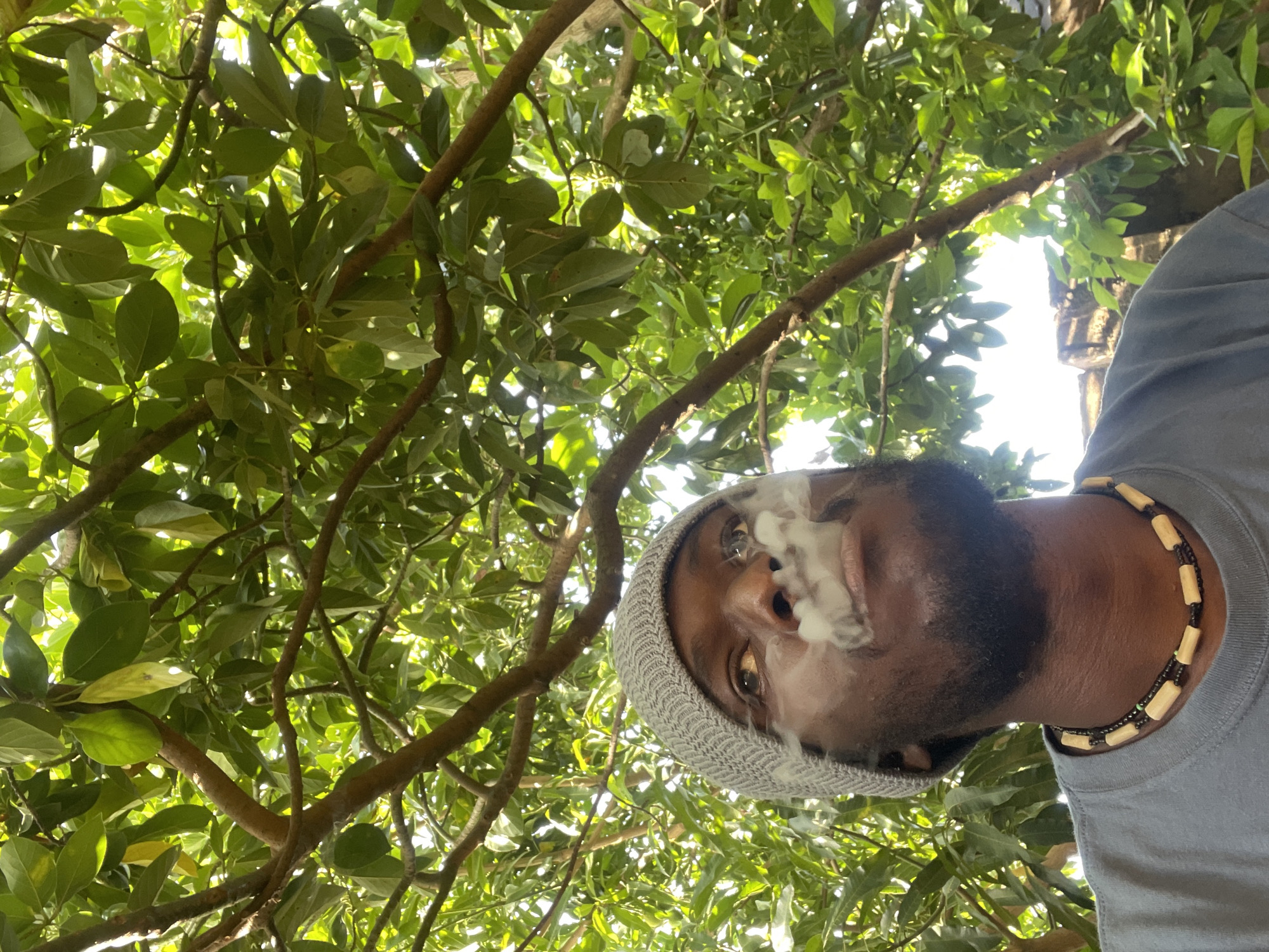 A low-angle medium shot shows a Black man exhaling smoke upward, with a green leafy canopy filling most of the frame. The man wears a grey knit cap and a two-strand necklace of tan and black beads. He has a dark beard. The location is possibly Jos, Nigeria, but this is not definitively verified by any visible elements in the image. The surrounding environment appears to be wooded.