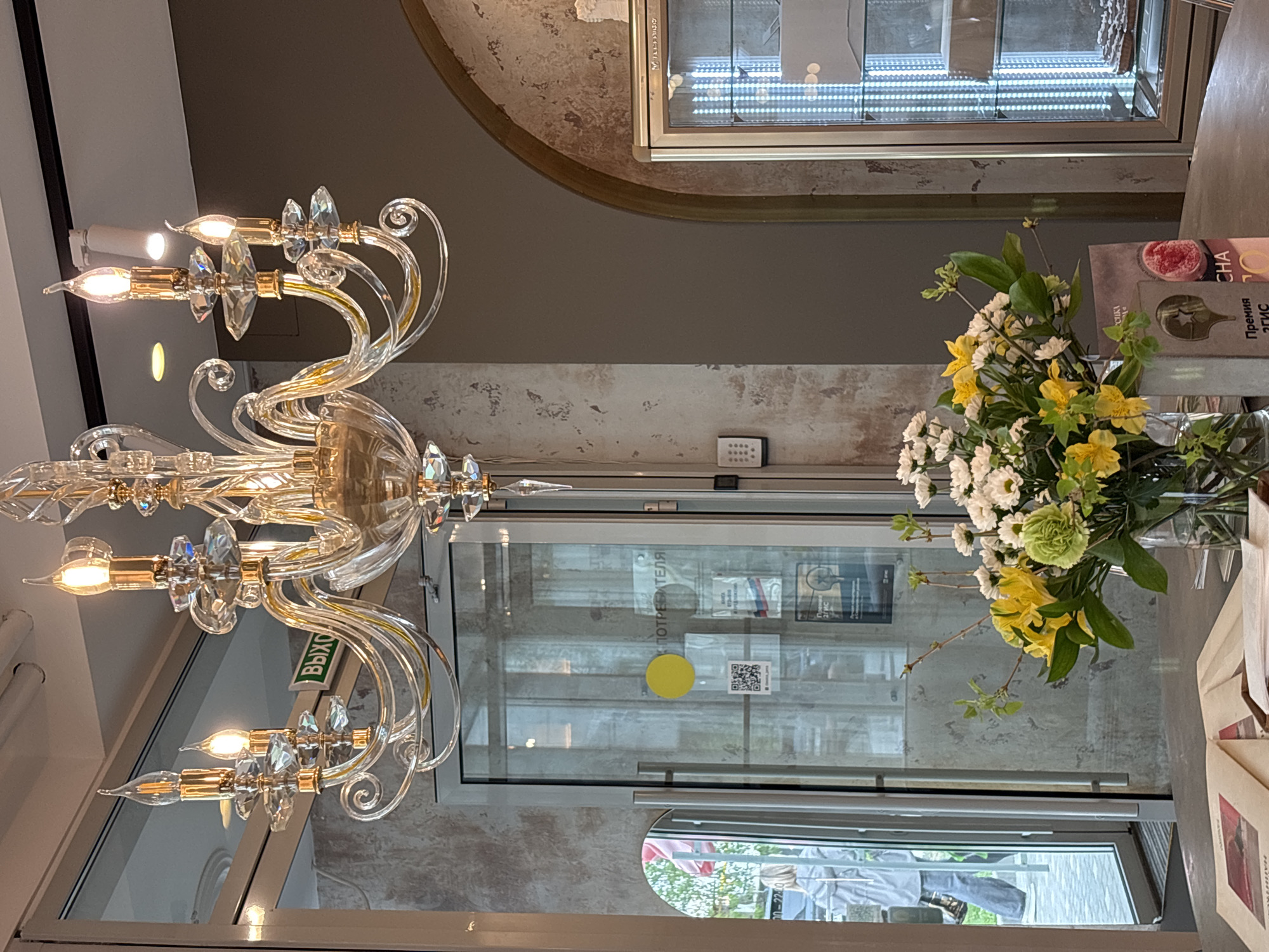 The image shows a view of a cafe in Perm, Russia. A large, ornate chandelier hangs in the foreground. A bouquet of yellow and white flowers sits on a table near a window. The window looks out onto a street scene. A refrigerated display case is visible in the background.