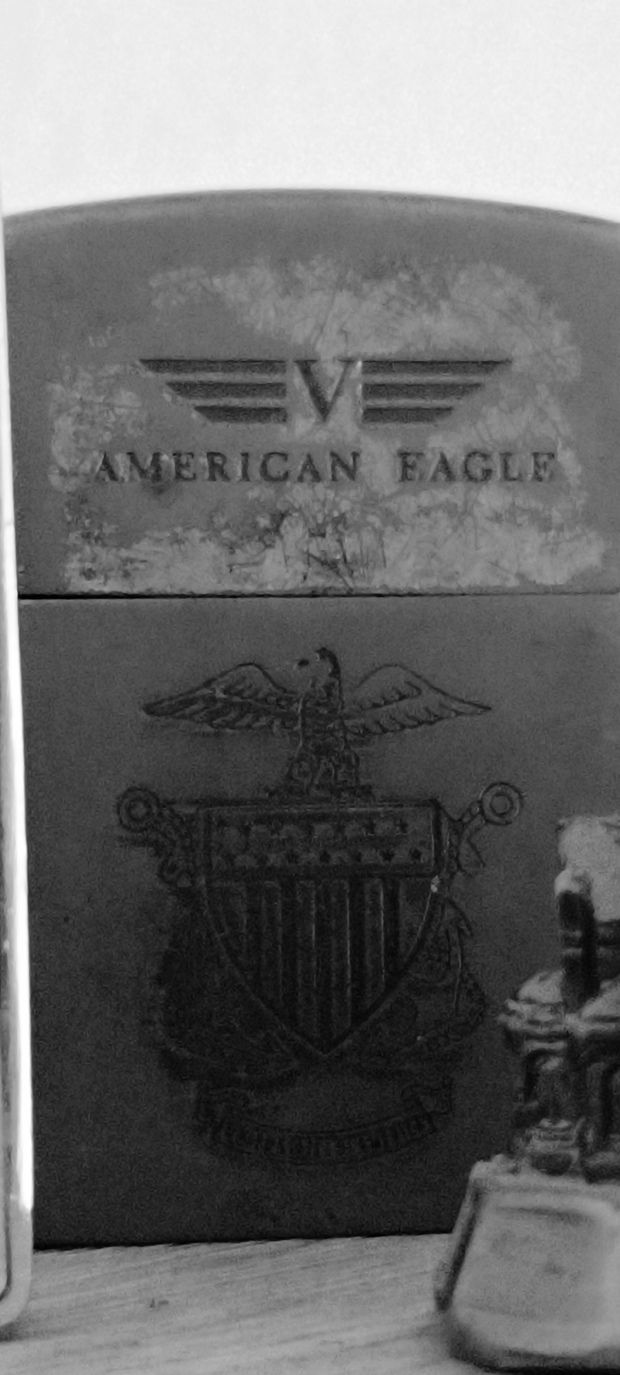 This is a grayscale, close-up, eye-level shot of an object that appears to be a lighter, with a metallic casing that has a worn and textured surface. The top section of the casing features an embossed logo with the words "AMERICAN EAGLE" below a stylized eagle or wing symbol. The bottom section has a more detailed embossed crest with an eagle perched atop a shield, flanked by decorative elements. The object rests on a surface that appears to be wood, with a subtle grain visible. To the right, a portion of another object, possibly a statue or figurine, is visible in the foreground, also showing signs of wear and texture. The overall impression is one of aged metal and craftsmanship. There is no indication of the location in Winnenden, Germany, or any people, actions, or interactions.