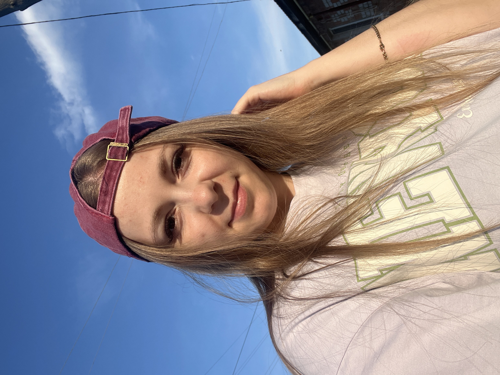 The image shows a young woman wearing a maroon baseball cap and a light purple t-shirt with green lettering. She is outdoors, with a bright blue sky in the background. The sun is casting a warm light on her face, and her hair is long and blonde. She is smiling and touching her hair with her right hand. Her left arm is visible, and she is wearing a delicate gold bracelet. Behind her, there is a hint of a building with a brick facade and a dark roofline. The scene suggests a casual, sunny day. The image was taken in the city of Mal'tsevo, Russia.